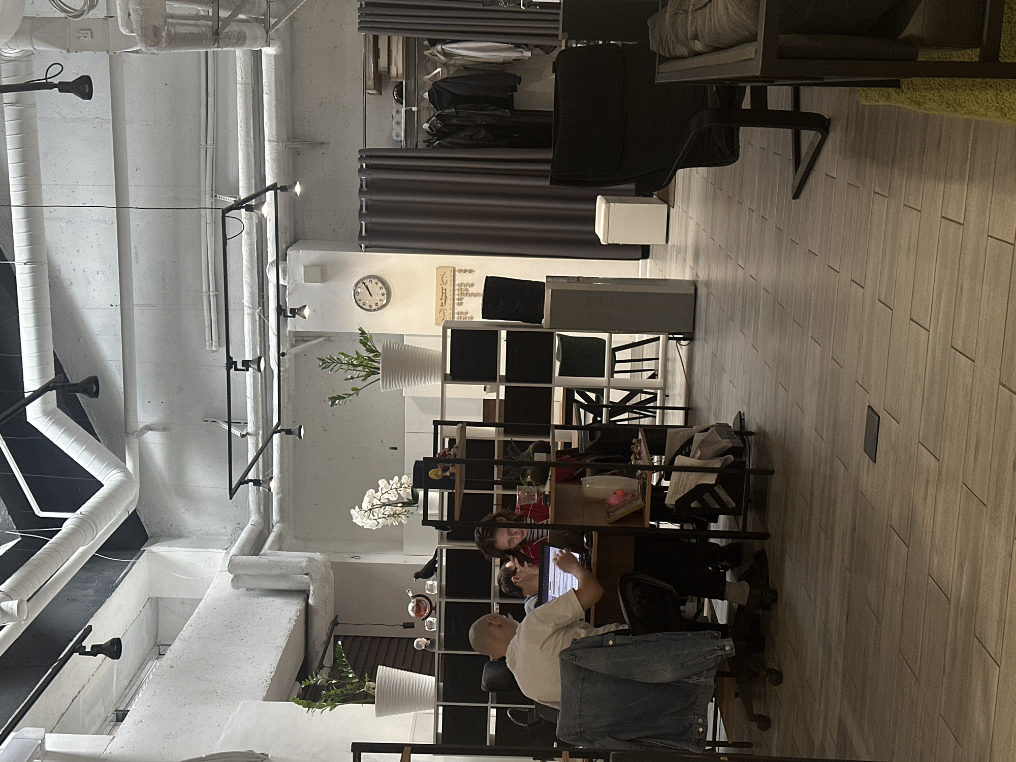 The image depicts an indoor coworking space, likely in Moscow, Russia, characterized by exposed industrial elements such as pipes and ventilation systems against a white, textured ceiling and walls. The floor is tiled with a wood-grain pattern.
In the foreground, a bald man in a light-colored t-shirt and dark pants is seated at a desk, facing away from the camera, and appears to be working on a laptop. A denim jacket is draped over the back of his chair. To his right, a woman with glasses and dark hair, wearing a red jacket and striped top, is also seated at the same desk, looking at the laptop. A second person is partially visible between them, also appearing to work at the computer.
Behind the desk, a shelving unit contains various items, including plants in white pots, decorative objects, and what appear to be figurines. A large white air purifier stands to the right of the shelving unit.
Further into the space, dark grey curtains hang, obscuring a section of the room. To the right of the curtains, a closet or dressing area is visible, with clothes hanging on racks. In the background, a clock on the wall shows approximately 4:10.
The overall atmosphere suggests a functional and somewhat utilitarian workspace, common in modern co-working environments.