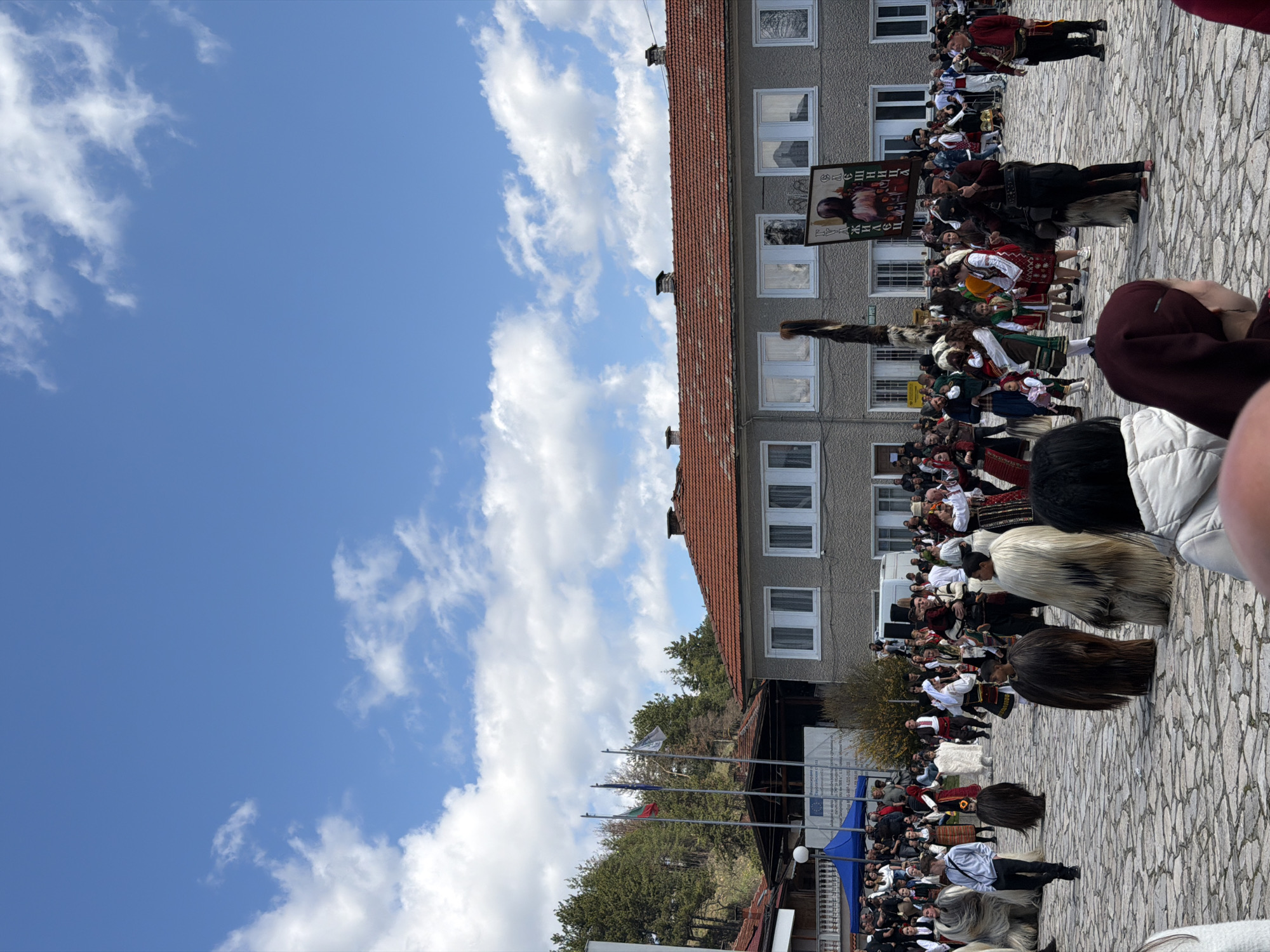 A vibrant cultural event is taking place in Eleshnitsa, Bulgaria. A crowd of people, many dressed in traditional Bulgarian folk costumes, has gathered on a cobblestone square in front of a multi-story building with a red-tiled roof. Flags of Bulgaria and the European Union are visible. Some individuals are wearing large, furry costumes, possibly related to a local festival or ritual. The sky is bright blue with scattered white clouds. The scene appears to be a public celebration or performance, with a significant number of participants and spectators.