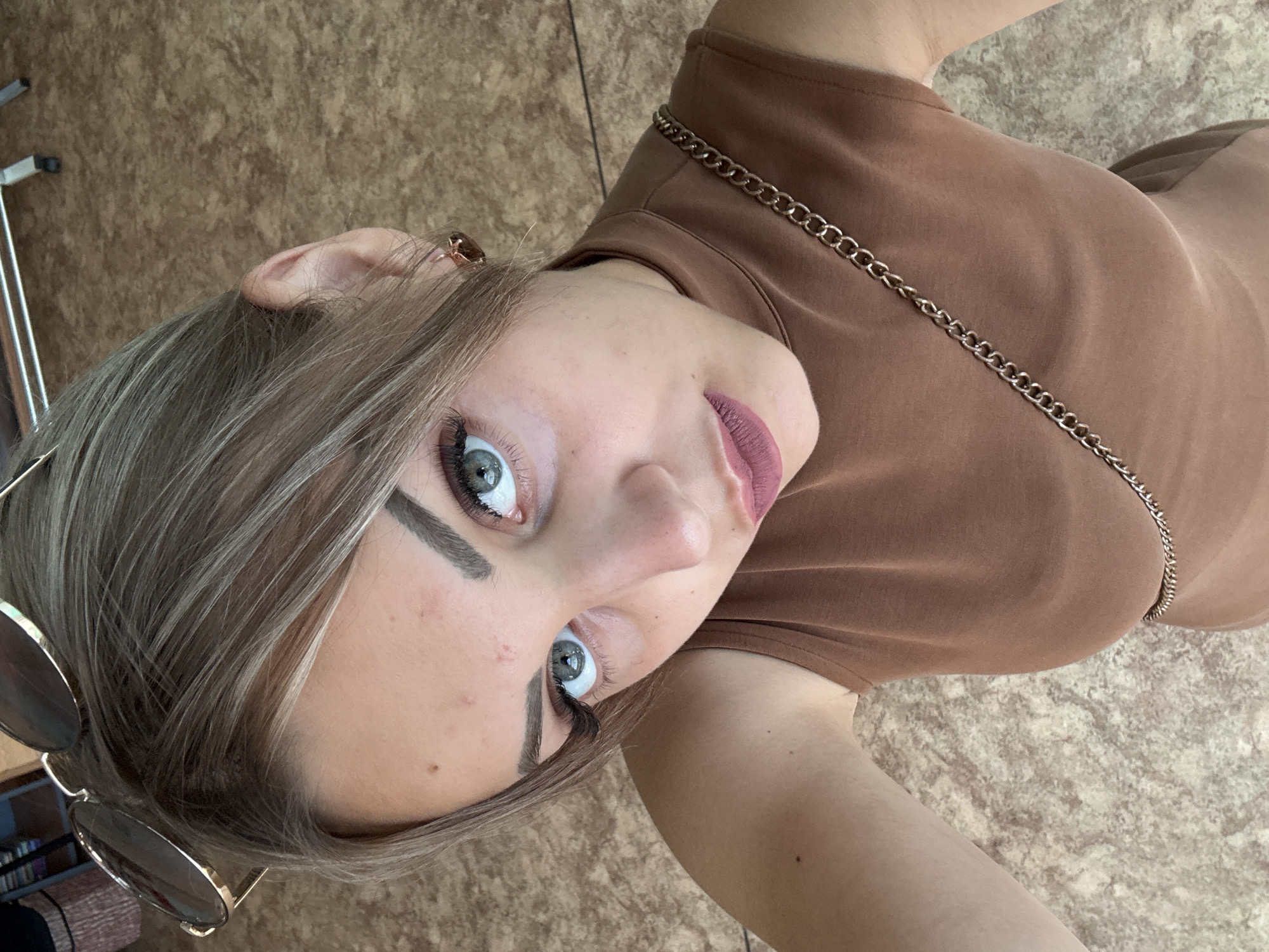 A woman with blonde hair and blue eyes is looking up at the camera. She is wearing a brown sleeveless top with a gold chain strap and sunglasses on her head. The background appears to be an indoor setting with a patterned floor. The image does not provide any information about the city of Yuzhno-Sakhalinsk, Russia.