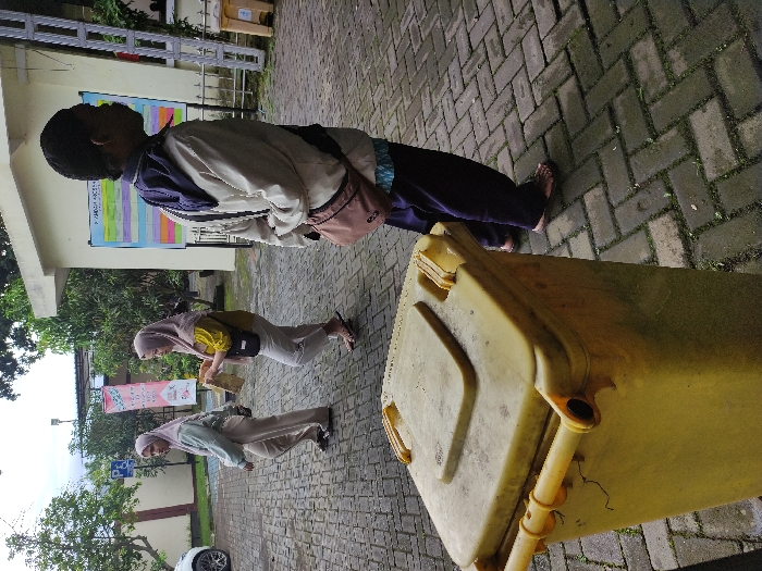 A slightly blurry outdoor photograph captures four individuals and a large yellow trash bin on a paved brick ground in what appears to be Kepanjen, Indonesia. The image is rotated counter-clockwise.

In the foreground, a large, faded yellow, wheeled trash bin occupies the right side of the frame. To its left, a person, likely male, stands facing away from the camera. This individual wears a light-colored jacket over a dark top, dark pants, and a brown shoulder bag, and appears to be barefoot.

Further back and to the left are two women, both wearing hijabs. One is dressed in a yellow top and light-colored pants, holding an object that resembles a book or folder. The other woman, slightly behind and to the left, wears a light green top and light pants. A fourth person, mostly obscured by the first individual, is partially visible.

The setting is an outdoor paved area, possibly a courtyard or walkway outside a building. The ground is made of grey interlocking bricks, with some moss visible in the cracks. In the background, a light-colored building wall features a large, colorful chart or poster with multiple columns and rows of text, largely unreadable but suggesting an informational display. Further behind the women, another banner or flag with some unreadable text is visible, alongside a blue sign with a white wheelchair symbol, likely indicating accessibility or parking. Trees and foliage are present on the left side of the frame, and a white car is partially visible in the far bottom left. The lighting suggests daytime, possibly overcast. The individuals appear to be standing casually.