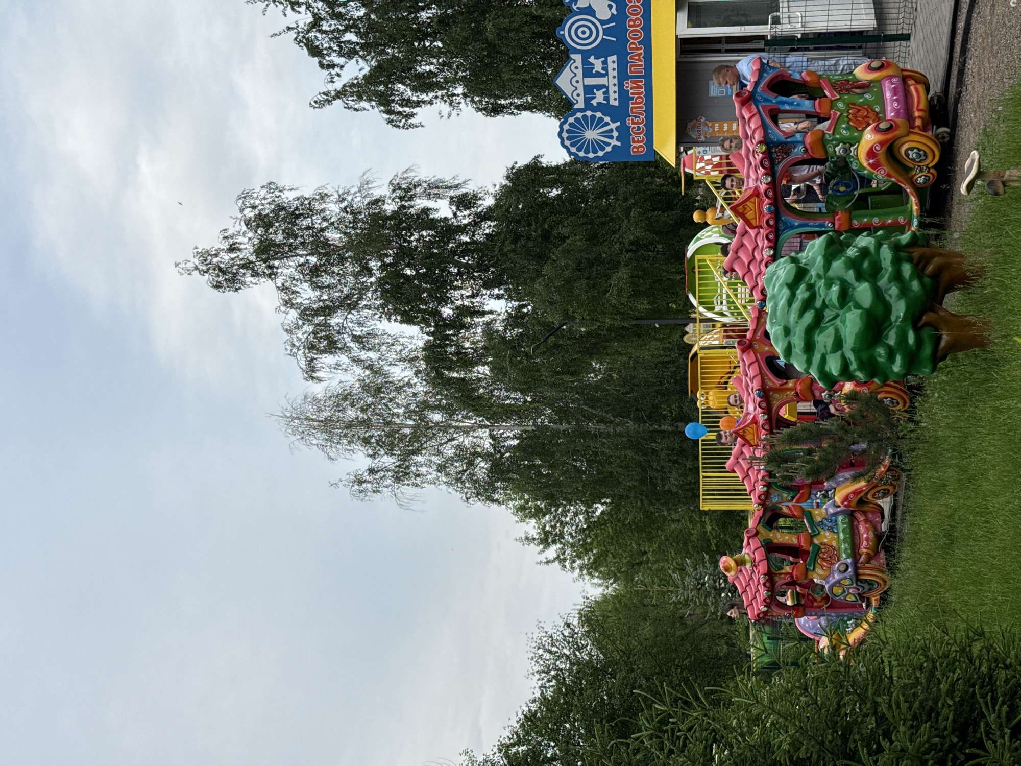 This image depicts a children's amusement park ride in Krasnoyarsk, Russia. The ride appears to be a colorful, cartoonish train with multiple cars, each decorated with vibrant colors and whimsical designs. Several people, including children and adults, are visible on the ride, either seated or standing.
In the background, there is a yellow fence surrounding the attraction, and a building with a blue sign that reads "ВЕСЁЛЫЙ ПАРОВОЗИК" (Vesyolyy parovozik), which translates to "Merry Little Train." The surrounding environment is characterized by lush green trees and grass, suggesting an outdoor setting within a park or recreational area. The sky is overcast, indicating a cloudy day. The overall scene captures a moment of enjoyment and leisure at a local amusement park.