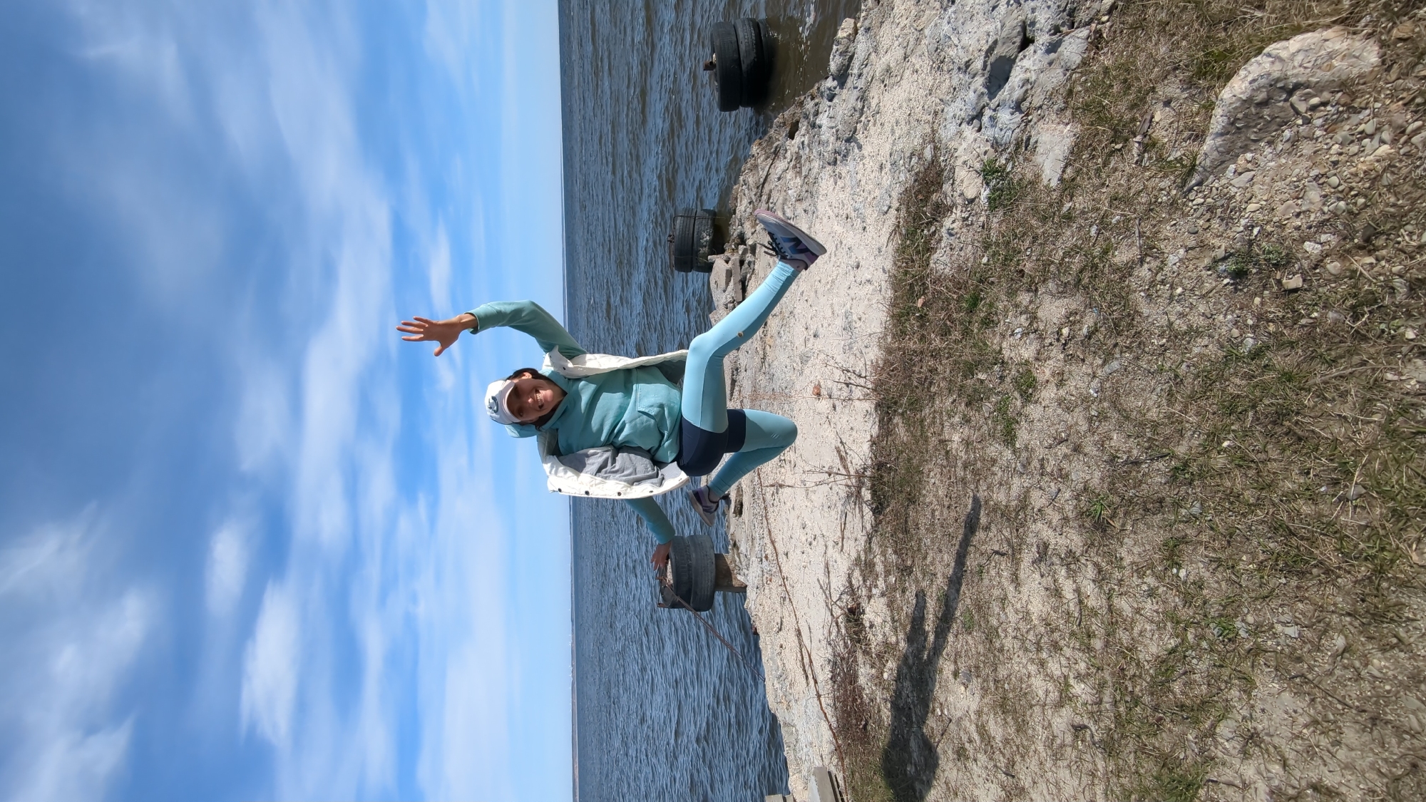A bright, outdoor scene depicts a person in a dynamic pose next to a body of water under a clear blue sky. The main subject is a woman, captured mid-action, possibly jumping or performing an acrobatic move. She is smiling broadly and looking towards the camera. She wears a light blue or teal hooded sweatshirt, matching light blue athletic leggings with darker blue shorts over them, a white sleeveless puffy vest, and a white baseball cap. Her athletic shoes are dark with light soles.
The setting features a calm, expansive body of water, likely a lake or sea, stretching towards the horizon. The water meets a rugged shoreline composed of rocks, sand, and patches of dry, sparse grass. Two large, black, discarded tires are partially submerged in the water near the shore. The sky above is a vibrant blue with scattered white cumulus clouds.
The bright, natural lighting indicates it is daytime, likely late morning or early afternoon, on a clear and pleasant day. The woman's joyful expression conveys a sense of happiness and playfulness. There is no visible text in the image.