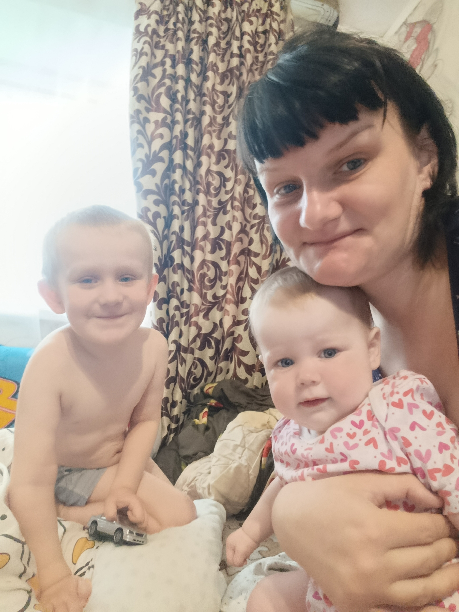 The image shows a woman with dark hair and bangs holding a baby in a pink outfit with red hearts. Next to them sits a young child with short hair and no shirt, holding a small toy car. They are all in a room with patterned curtains and bedding. The location is not specified.