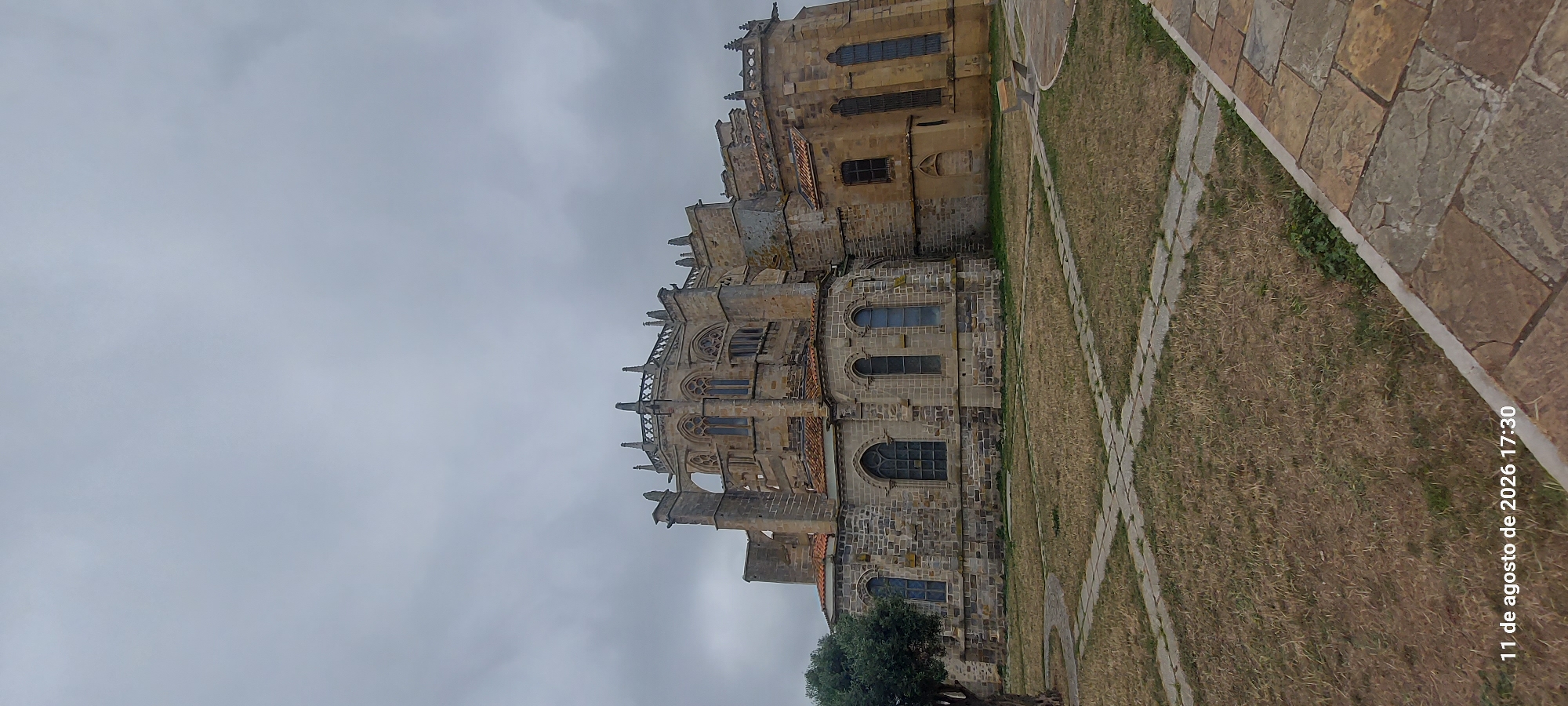 The image shows a large, gothic-style cathedral made of stone, with intricate architectural details and arched windows. The sky is overcast and grey. In the foreground, there is a lawn with a path made of stone slabs. A portion of a tree is visible on the left side of the image. The timestamp "11 de agosto de 2026 17:30" is visible in the bottom left corner of the image. The cathedral is identified as the Church of Santa María de la Asunción in Castro-Urdiales, Spain.
