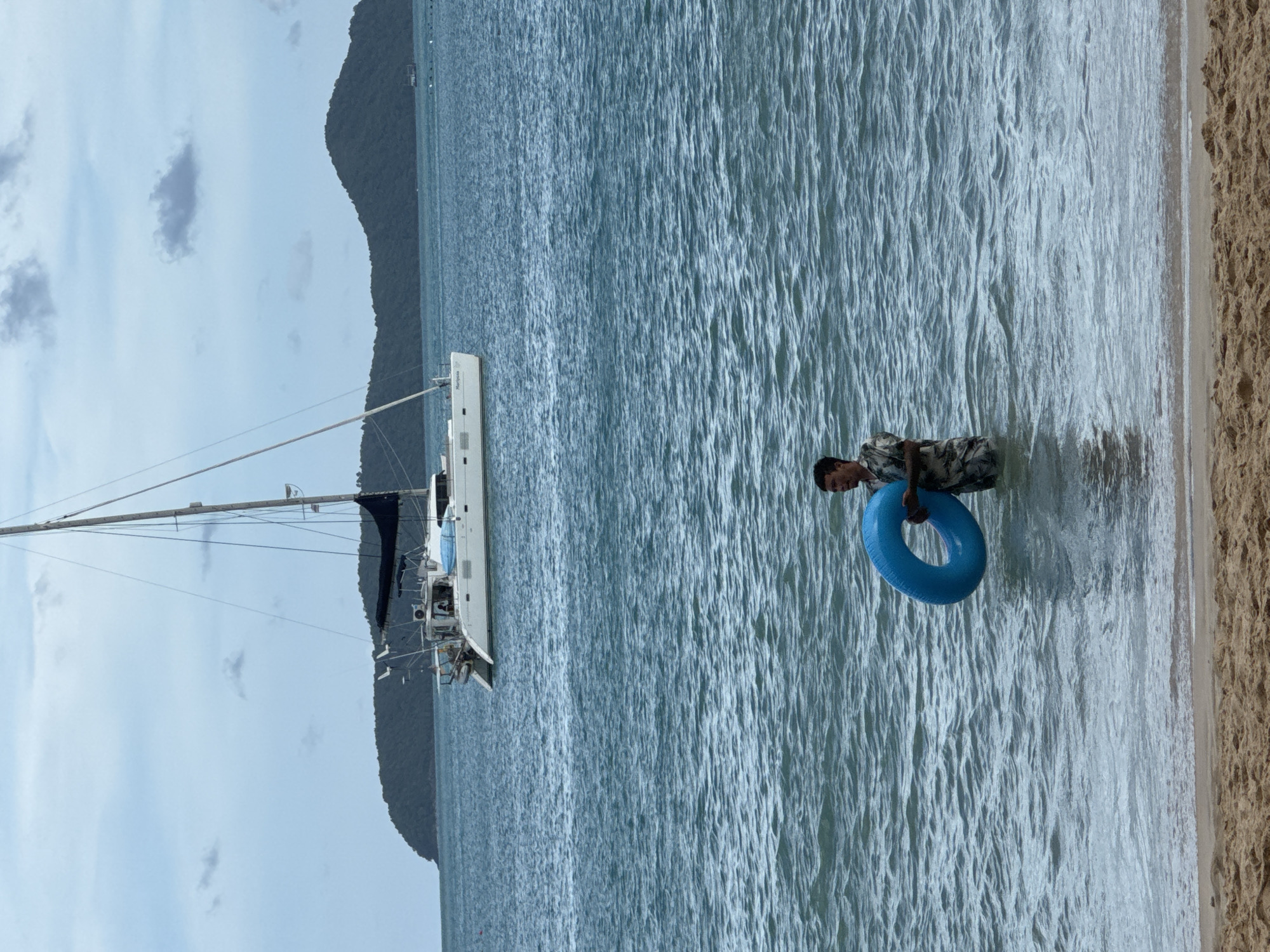 A man stands in the ocean with a blue inner tube, facing to the right. He is wearing a patterned shirt and pants. In the background, a white catamaran is anchored on the water. Behind the catamaran, a green, hilly island is visible under a cloudy sky. The scene appears to be a beach in Wichit, Thailand.