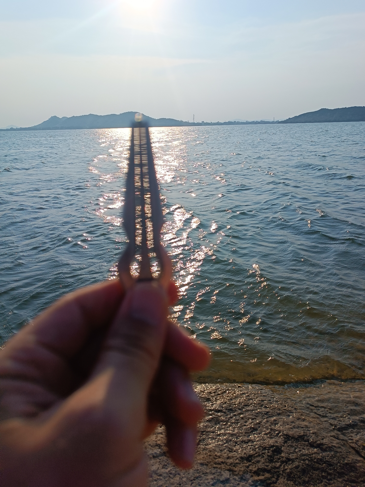 A handheld mouth harp is held up to the light of the sun in front of a body of water with a shoreline. In the distance are low-lying hills and an industrial structure. The location is Kammavaripalli, India. The subject is a single hand holding a mouth harp. The environment includes water, land, sky, and distant hills.