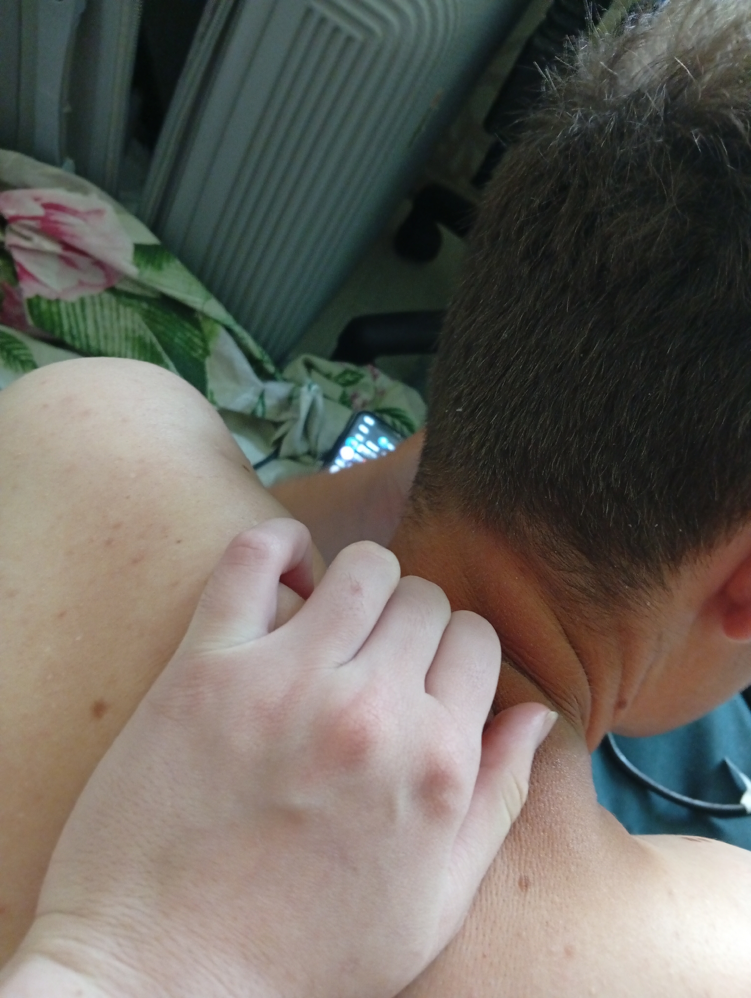 A close-up shot presents a person's neck and part of their head, with a hand massaging or gripping the neck. The person has short, dark hair and is facing away from the camera. A light-skinned hand is applying pressure to the back of the neck. A floral patterned fabric and a white slatted object, possibly a radiator or air conditioner, are visible in the background. A phone with its screen illuminated is visible. Based on the content alone, there is no indication of the location being Poltava, Ukraine.