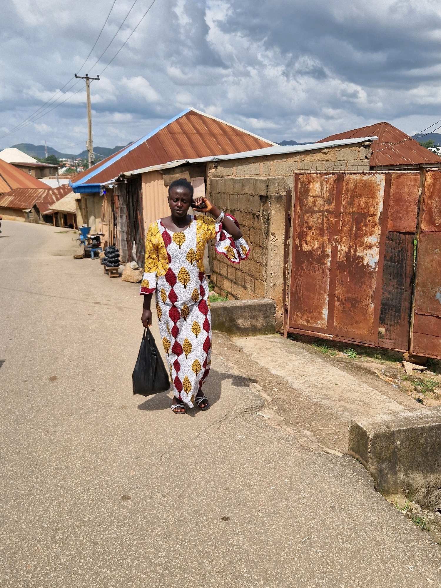 A medium-shot, eye-level, full-body outdoor photograph captures a woman walking down a street in Kabong, Nigeria. She is positioned centrally in the frame, facing forward and slightly to the left of the viewer. The woman is wearing a long, colorful dress with a white base adorned with a repeating pattern of red and yellow leaf-like shapes. Her left hand holds a black plastic bag, and her right hand is raised to her ear, as if holding a phone. She is wearing open-toed sandals.
The environment is a paved street with a rough, speckled surface. To the left of the woman, the street recedes into the distance, lined with low-rise buildings that have corrugated metal roofs, some showing signs of rust. A wooden utility pole with overhead wires stands on the left side of the street. To the right of the woman, there is a section of brick wall and a large, rusty, corrugated metal gate. The ground to the right of the gate is a mix of concrete and dirt, with some sparse vegetation. In the background, more buildings with similar corrugated roofs are visible, and the landscape suggests a hilly terrain. The sky is partly cloudy with large, grey clouds dominating the upper portion of the image. The overall lighting indicates it is daytime.