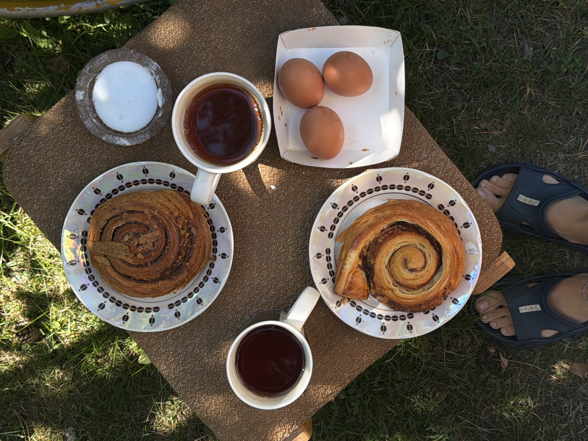 An outdoor picnic is laid out on a brown, textured placemat on a grassy area, likely in Udelnaya, Russia, given the context. The arrangement includes two spiral pastries, one on a decorative white plate in the upper left and another on a similar plate in the lower center. Adjacent to the upper pastry is a small, circular stone dish filled with white granular material, possibly sugar. Two white mugs filled with dark liquid, presumably tea or coffee, are positioned on the placemat, one to the left of the upper pastry and the other to the right. To the right of the mugs and lower pastry is a small white cardboard container holding three brown eggs. The scene is viewed from directly overhead, with a person's feet wearing dark blue sandals visible at the bottom of the frame. Sunlight casts shadows across the grass and the placemat, suggesting it is a bright day.