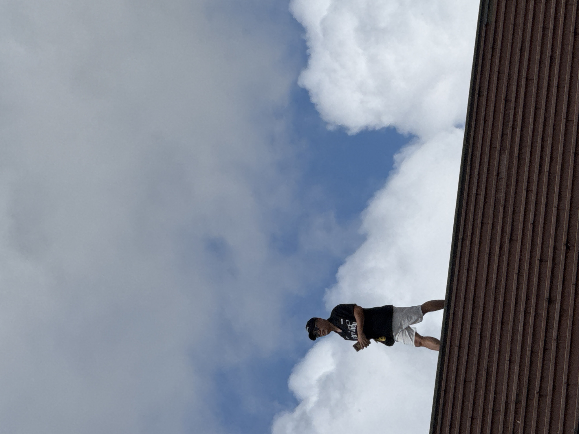 A man in a black polo shirt and white shorts stands on a brown tiled roof, looking to his left. He is wearing sunglasses and a black baseball cap. The sky is filled with white clouds against a blue backdrop. The image was captured in the city of Da Lat, Vietnam.