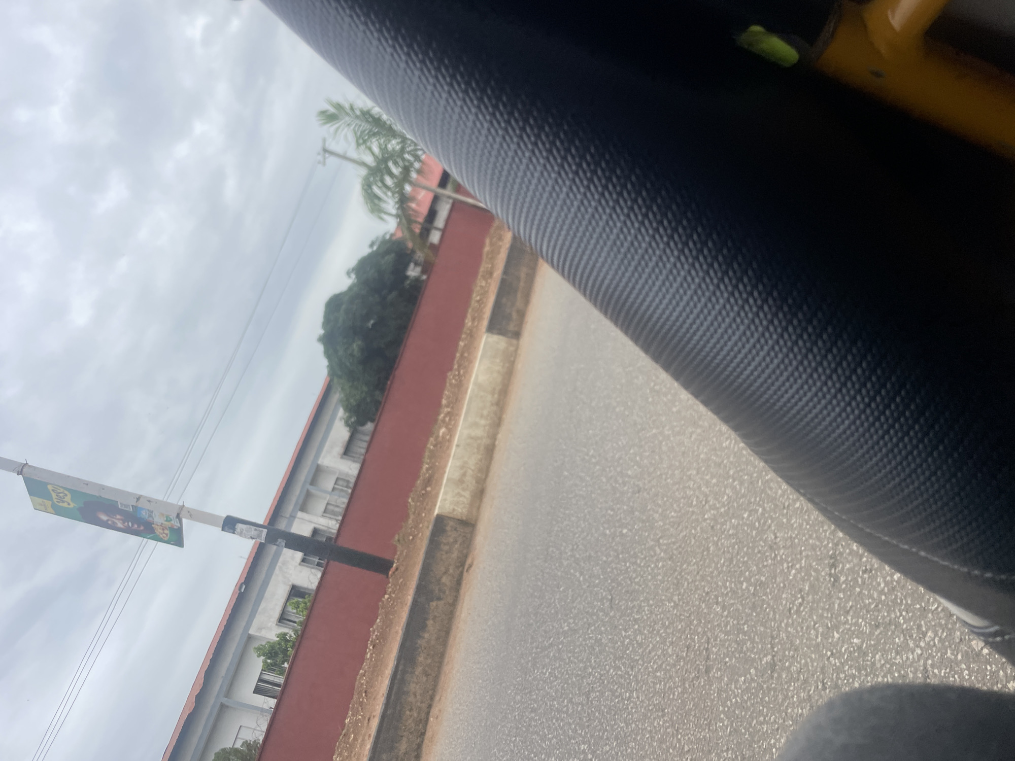 A tilted, full shot shows a street scene in Jos, Nigeria, under an overcast sky. In the foreground, a black textured surface, possibly a vehicle seat, dominates the right side of the frame. Beyond it, a paved road stretches into the distance, with a greyish-brown curb bordering a reddish-brown wall. The wall encloses a building with a light-colored facade, visible behind it, with barred windows and a few green trees. To the left of the wall, a tall pole stands, adorned with a green and yellow advertisement featuring a woman's face and text. Further back, palm trees and lush green foliage can be seen. The overall impression is that of an urban environment, possibly on a cloudy day.