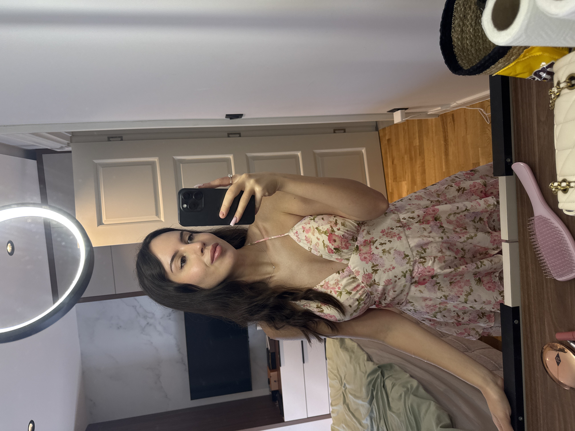 A young woman with long, dark hair is taking a mirror selfie in a bedroom. She is wearing a light-colored, floral-print dress with spaghetti straps. She is holding a black smartphone in her right hand, which is capturing the reflection. Her left arm is resting on a dark surface in front of the mirror. The bedroom appears to be well-lit, with a large ring light visible above the mirror. In the reflection, a bed with a green comforter is visible on the left, and a white dresser with a television on top is in the background. A doorway and part of a wall are also visible. On the surface in front of the mirror, there are various items, including a pink hairbrush, a lipstick, a compact makeup case, and a white quilted handbag with gold chain straps. A woven basket with a yellow bag and toilet paper rolls is also present. The floor is made of polished wood.