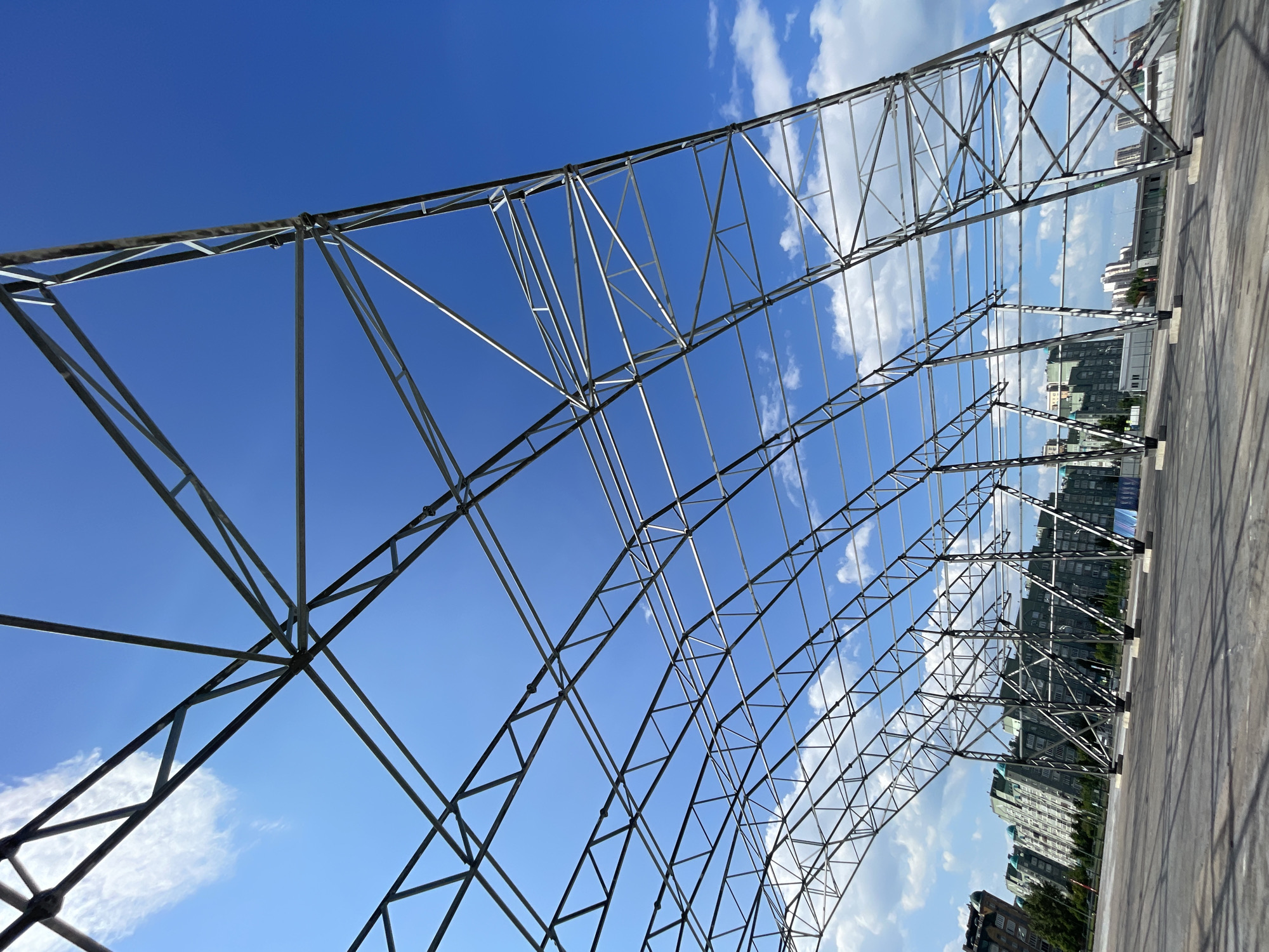 The image shows the partially constructed steel frame of a large-scale, arched structure against a bright blue sky with scattered white clouds. The structure is composed of a complex network of intersecting metal beams and trusses, creating a geometric pattern. In the background, several multi-story buildings with modern architecture and green accents are visible, suggesting an urban or suburban setting. Trees line the street in front of the buildings. The ground in the foreground is a paved, flat surface, likely a plaza or an open area. There are no people visible in the image. The scene appears to be an outdoor construction site. The presence of the specific building styles and the general urban landscape suggest the location could be Kazan, Russia.