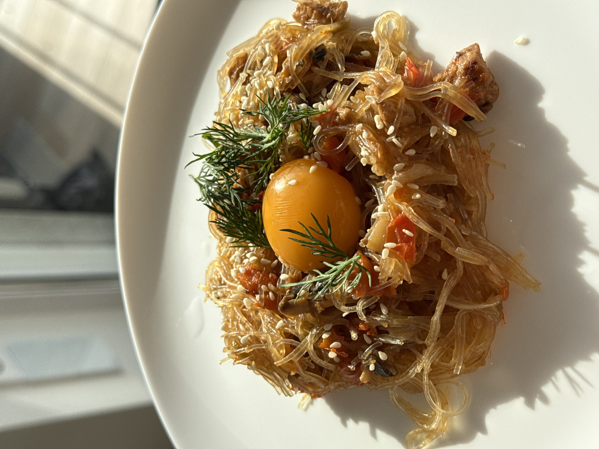 A top-down shot displays a plate of Asian-inspired cuisine, likely served in Perm, Russia. The primary dish appears to be translucent glass noodles tossed with vegetables, possibly tomatoes and mushrooms, and small pieces of meat. The dish is generously sprinkled with sesame seeds. At the center is a single egg yolk surrounded by sprigs of dill. The food sits on a white plate, illuminated by strong sunlight casting shadows to the right. A portion of a window is visible on the left.