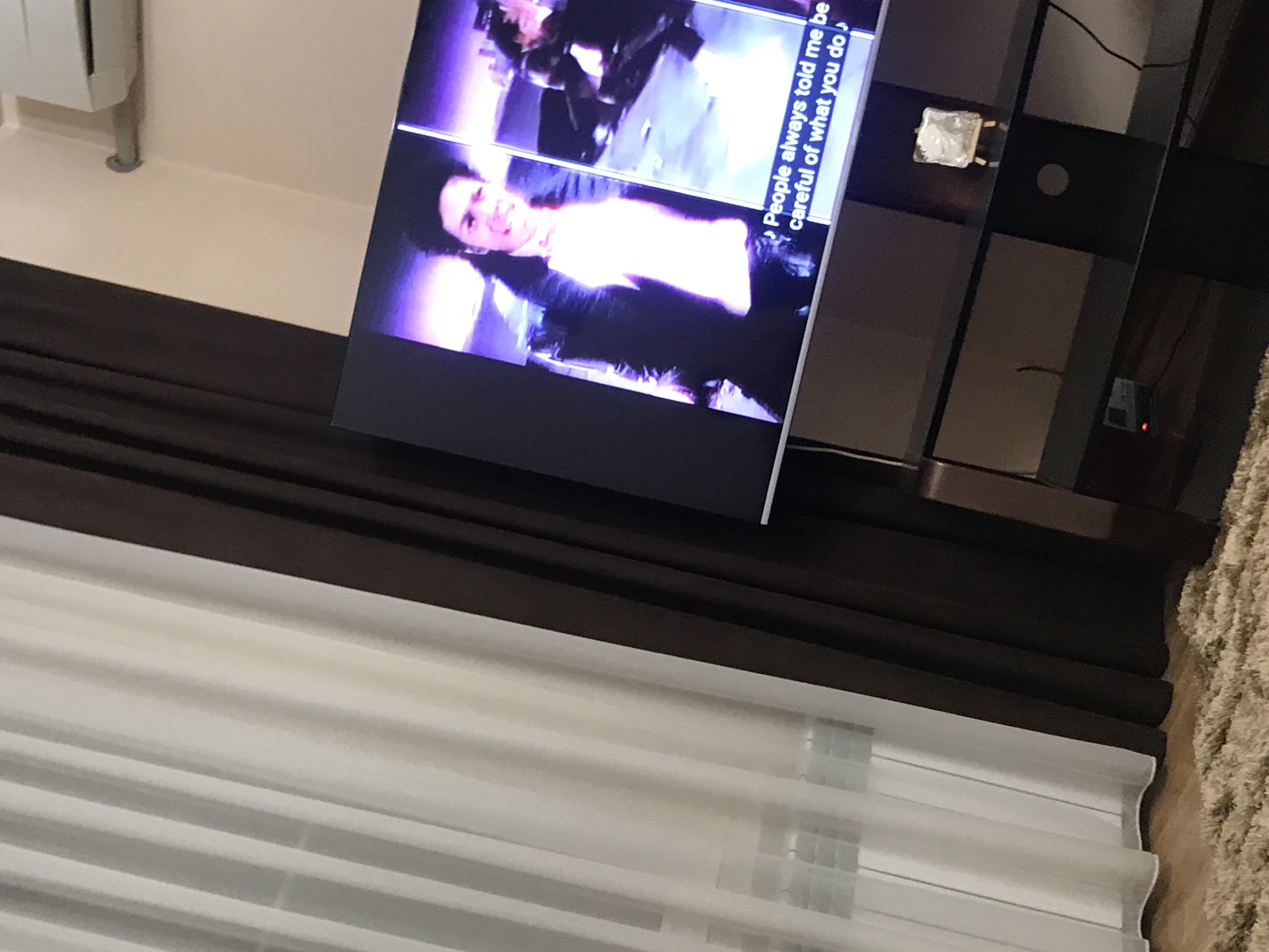 The image depicts a television screen displaying a scene with a person on a stand in what appears to be a living room. The TV is situated on a dark brown multi-tiered stand. In front of the stand and partially obscuring the view, there are white, pleated blinds. Above the blinds are dark brown curtains. To the side of the stand there is an off-white wall. A brown carpet partially covers the floor. The specific location in Yerevan, Armenia, cannot be determined from the image.