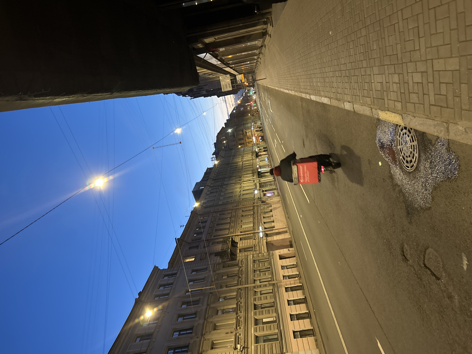 The image depicts a street scene viewed from a high angle, oriented vertically. The scene is a city street with buildings lining one side and a sidewalk on the other. A person on a scooter, distinguished by a red delivery bag on their back, is moving along the street. The sky is a deep blue, suggesting either early morning or late evening. Streetlights are visible, casting a warm glow on the pavement. The buildings appear to be old, with many windows and architectural details. The street is paved with asphalt, and the sidewalk is made of bricks. There is a manhole cover on the sidewalk, and a small patch of what looks like scattered gravel or debris near it. The overall impression is one of a quiet, urban environment at dusk or dawn. There is no discernible text in the image.