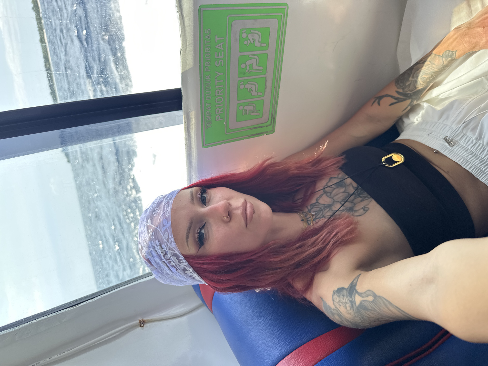 A medium shot captures a woman with reddish-brown hair, a lace headscarf, and several tattoos on her arms and chest. She is lying against a seat with blue, red, and white upholstery. She is wearing a black top and white shorts. Behind her, a sign indicates "PRIORITY SEAT" in Indonesian and English, accompanied by pictograms. A window shows a view of water, suggesting the woman is on a boat. The location is Unknown, Indonesia.