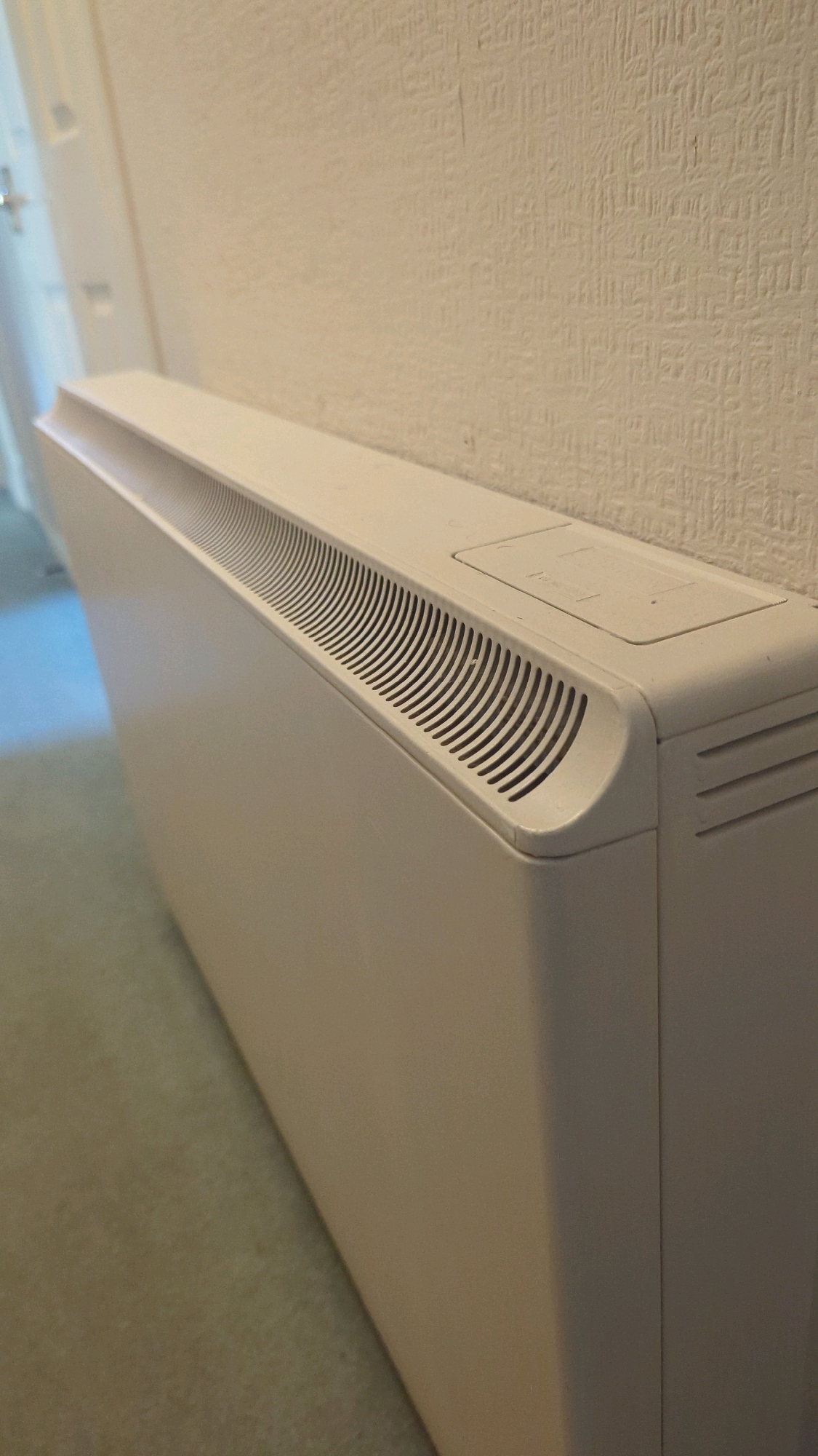 The image shows a close-up, angled view of a cream-colored electric convector heater mounted on a wall. The heater is rectangular with a ribbed ventilation grill along its top front edge. A small, rectangular panel is visible on the top surface of the heater towards the right. The wall behind the heater has a textured, off-white wallpaper. To the left of the heater, a section of a cream-colored door frame with a door handle is visible, and the floor appears to be carpeted in a light brown or beige color. The scene is indoors, suggesting a residential or commercial interior. The image does not depict any people or specific activities. The location is stated to be Cornelscourt, Ireland, but this information is not visually verifiable from the image itself.