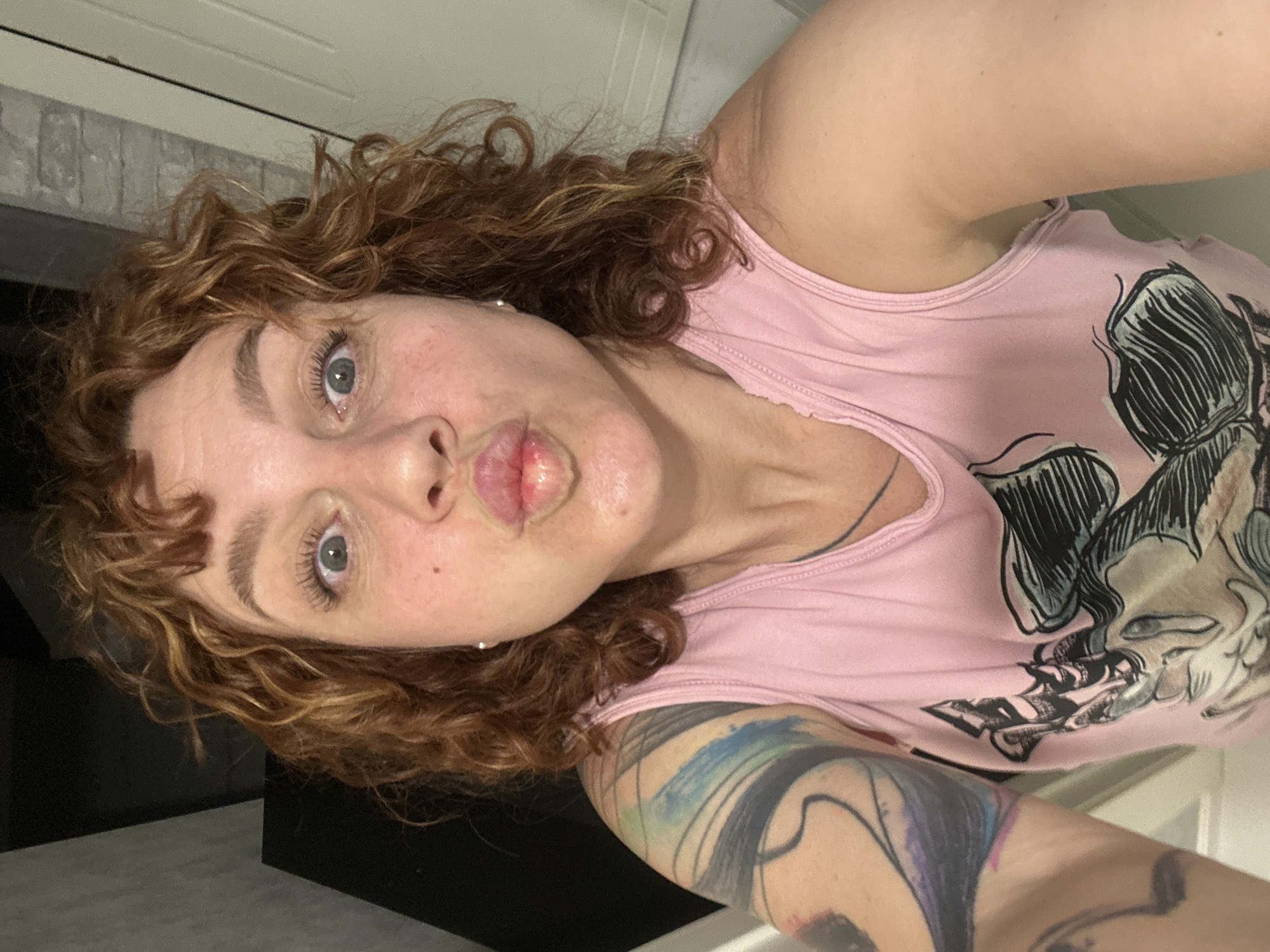 A woman with curly auburn hair and light eyes poses for a selfie. She is wearing a light pink tank top with a black and white graphic design. Her left arm, visible in the foreground, is adorned with a colorful tattoo. She is making a kissing face, and the background includes a brick wall and a dark surface. The location is not discernible as Sochi, Russia, from this image.