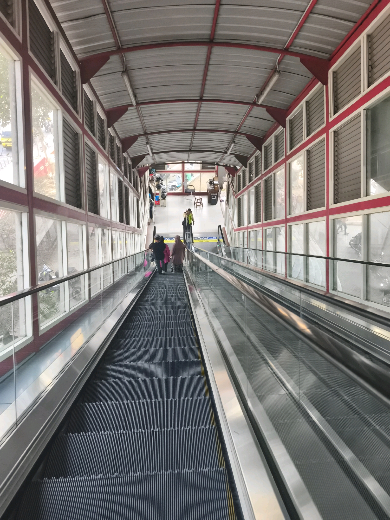 The image captures an escalator in what appears to be a covered pedestrian walkway or bridge, possibly at a transit station, located in Kelurahan 16 Ilir, Kecamatan Ilir Timur I, Indonesia. The escalator, seen from a top-down perspective, is angled downwards and has dark treads with yellow warning strips. On the escalator, three individuals are visible: two people in the foreground walking down, one wearing a dark top and the other in a light pink hijab, and a third person further up, partially obscured, also moving downwards. On the right side of the escalator, a person in a bright yellow jacket is standing, possibly attending to the escalator or overseeing the area. The walkway features a metal roof structure supported by red beams and has large glass windows on both sides, offering glimpses of the outside environment, including trees, a yellow vehicle, and parked motorcycles. The overall environment suggests a public space designed for pedestrian movement, likely in a busy urban setting.