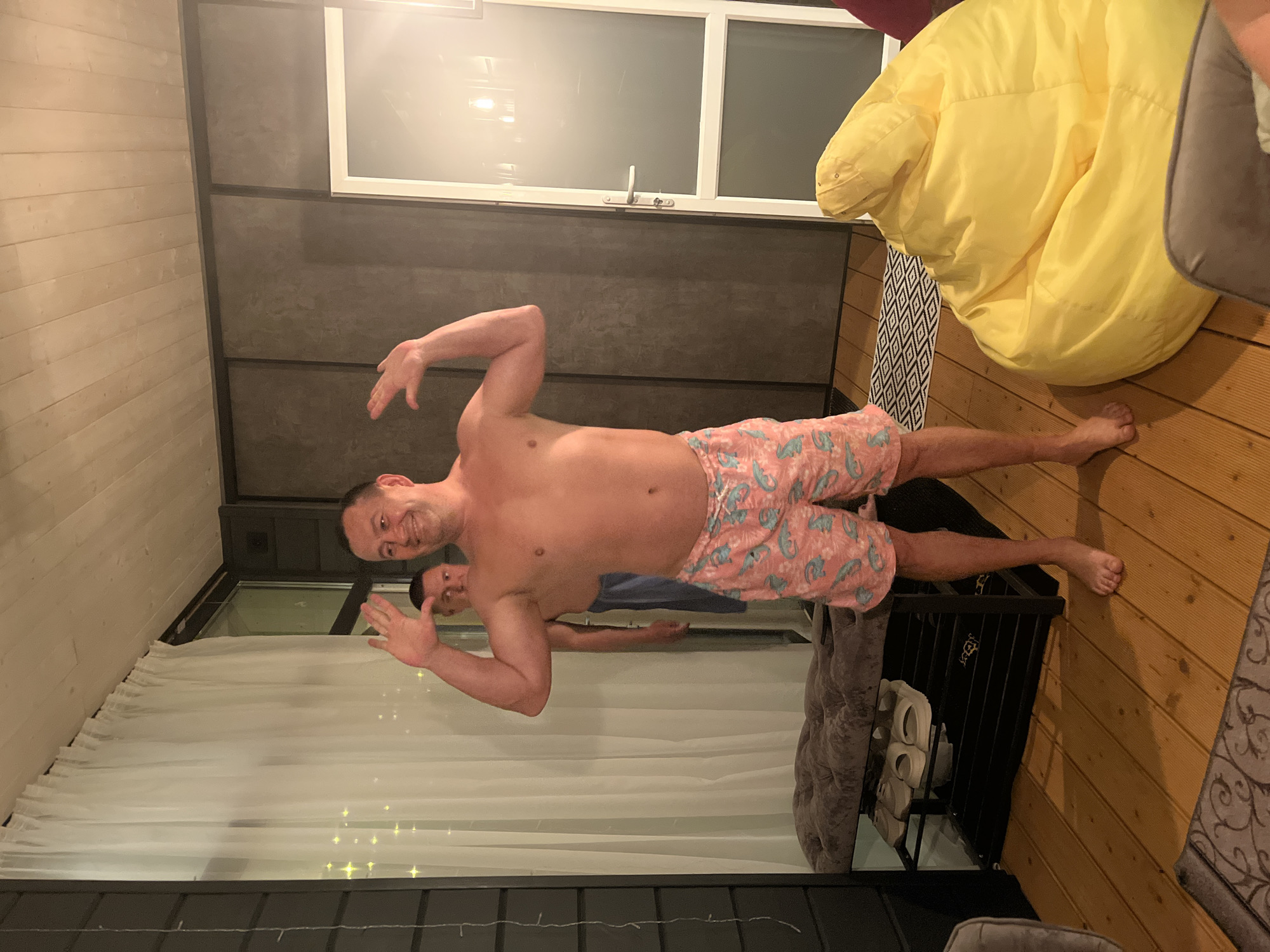 The image shows two men indoors. The man in the foreground is shirtless and wearing pink shorts with a blue dinosaur pattern. He is smiling and has his arms raised. A second man is partially visible behind him, wearing a blue shirt. The setting appears to be a living room, featuring a large window with sheer curtains, a beanbag chair, a rug, and a wooden floor. A shoe rack is partially visible near the center. The location is given as Новая Чернушка, Russia.