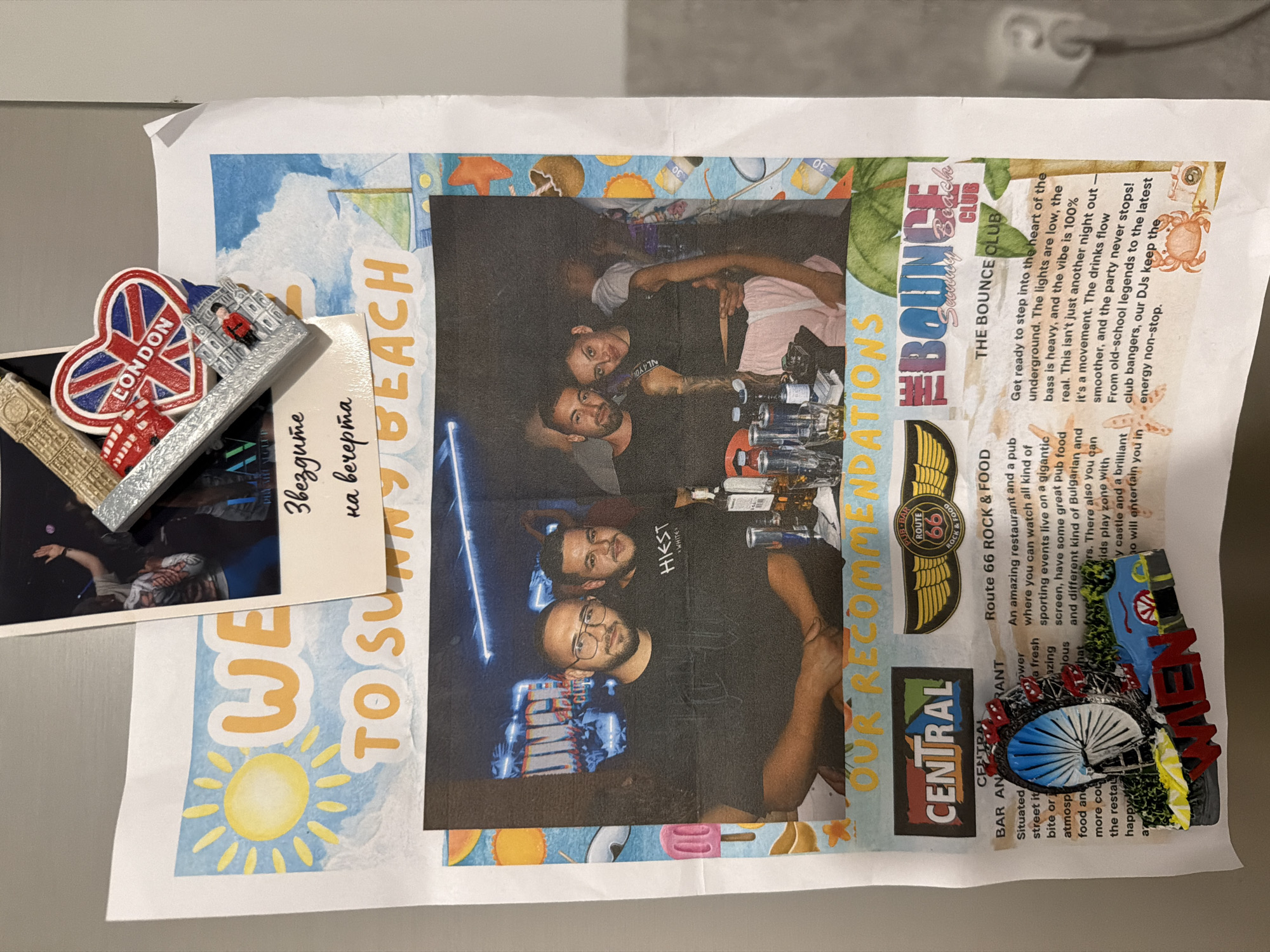 The image displays a collection of printed materials affixed to a metallic surface, likely a refrigerator. At the top, a portion of a photograph shows people at a concert or event, with a London-themed novelty item, featuring a red double-decker bus, a heart-shaped Union Jack, and the words "LONDON," positioned over it. Below this, a flyer or advertisement contains the text "WE TO SUNNY BEACH" in large, colorful lettering, accompanied by graphics of a sun and a sailboat.
The central focus is a newspaper clipping or magazine spread featuring two photographs. The first photograph shows three men, two in the foreground and one blurred in the background. The man on the left wears glasses and a dark t-shirt, while the man next to him wears a t-shirt with "HIKSST WHITE" printed on it. The second photograph captures a couple, a man with his arm around a woman, who is wearing a black crop top and pink pants. Both are looking towards the camera.
Below these photographs, the text "OUR RECOMMENDATIONS" is prominently displayed. To the left of this text is a section for "CENTRAL BAR AND RESTAURANT," with a graphic of a Ferris wheel and the word "WIEN" (Vienna) below it. To the right, there is a "ROUTE 66 ROCK & FOOD" advertisement with a graphic of wings and the Route 66 logo. Further to the right, an advertisement for "THE BOUNCE CLUB" on Sunny Beach is visible, with descriptive text about its atmosphere.
The image does not contain any information that would suggest a location in the city of Plovdiv, Bulgaria. The text and visuals explicitly refer to Sunny Beach and Vienna.