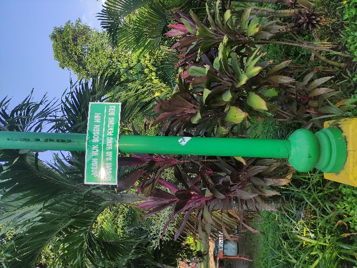 The image shows a green lamppost with a sign attached to it. The sign says "Jalan Yuk Bosen Nih! Laporan Aja Yang Penting Ber2." The lamppost is located in a park setting with lush green grass and various trees. The lamppost is located in Mojokerto, Indonesia, at the coordinates -7.464 latitude, 112.432 longitude. There are no people visible in the image.