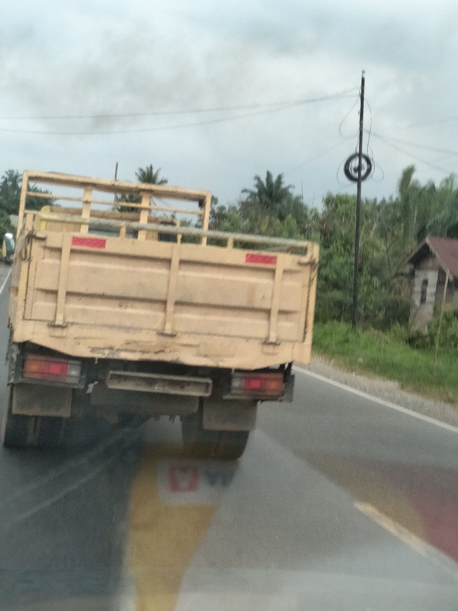 A faded, rear-view image captured from a vehicle shows a yellow flatbed truck driving on a paved road. The truck's bed is empty and its taillights are visible. The road has a white line marking its edge. To the right of the road, there is a grassy area with green foliage and trees. A small, weathered building with a dark roof is situated in the background, partially obscured by the trees. Overhead, a utility pole with a circular object attached stands against a cloudy sky. The image appears to have been taken in a rural or semi-rural area, consistent with the location of Bukit Selamat, Indonesia, given the presence of tropical vegetation and the type of building. The overall lighting suggests an overcast day.