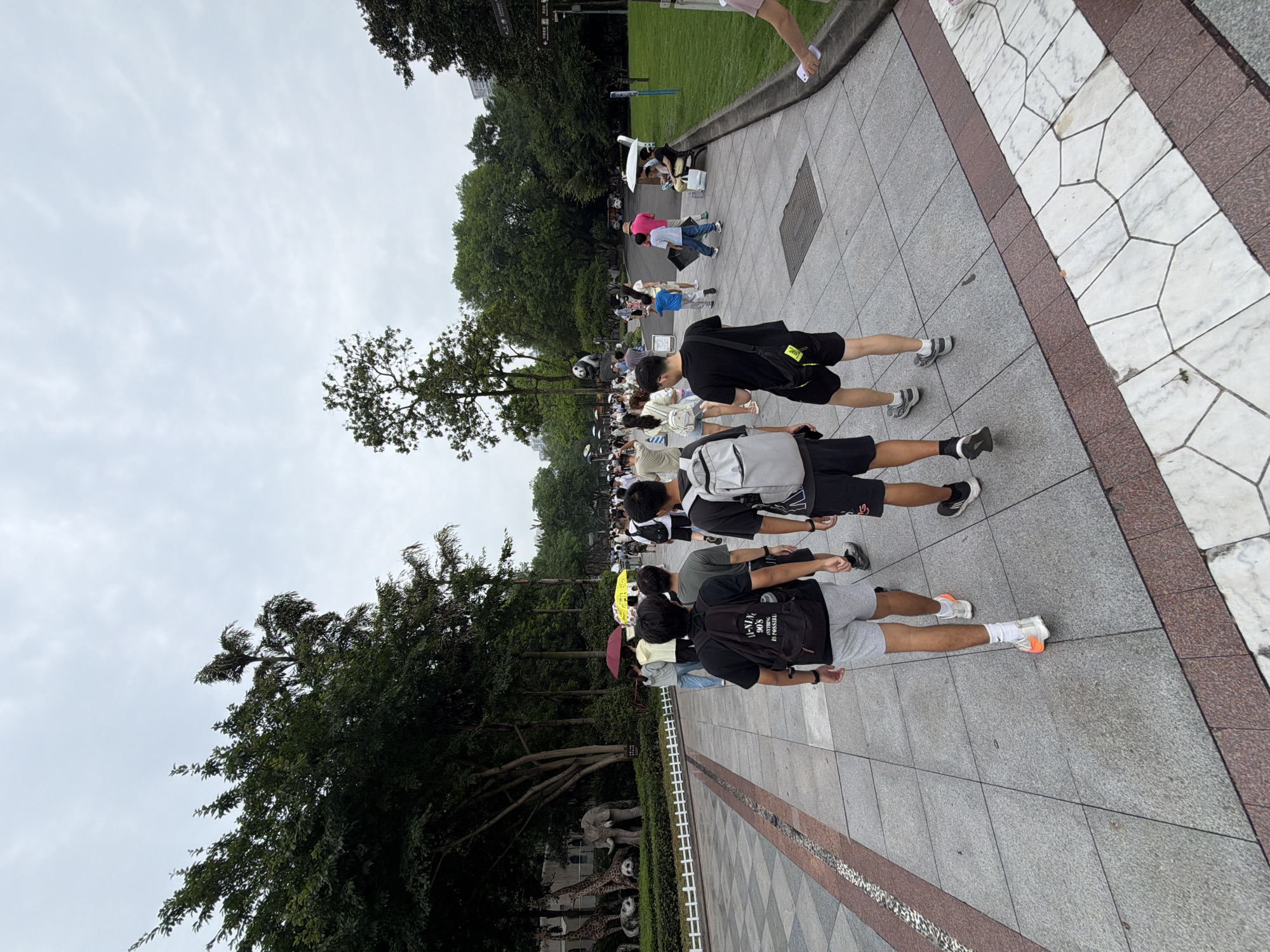 The image depicts a group of people walking along a paved pathway in an outdoor environment, likely a park or zoo, in Jiulongpo District, China. In the foreground, three young men are walking away from the camera, two of whom are wearing backpacks. The man on the left wears a black backpack with "VANS" printed on it and grey shorts. The man in the middle wears a grey backpack and black shorts. The man on the right wears a black backpack and black shorts. Further down the path, more individuals are seen walking, some with umbrellas. To the left of the pathway, there is a decorative display of animal statues including giraffes, an elephant, and pandas, with lush green trees and foliage in the background. On the right side of the pathway, a grassy area and more trees are visible, along with some benches. The sky is overcast.