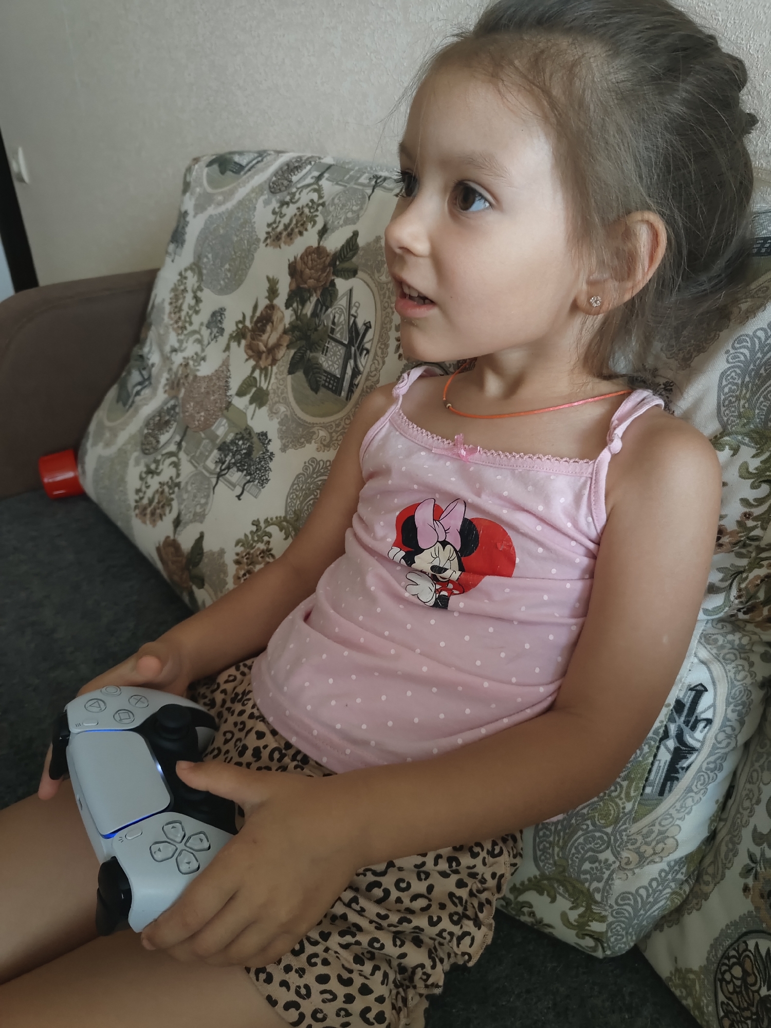 A young girl with brown hair and brown eyes is sitting on a couch, holding a white video game controller. She is wearing a pink tank top with white polka dots and a Minnie Mouse graphic on the front, and leopard-print shorts. A thin orange necklace is around her neck. She is looking off to the right side of the frame with an engaged expression. The couch has a patterned pillow behind her. A red object is visible on the left edge of the frame, behind the girl. The lighting suggests it is daytime. The setting appears to be indoors.