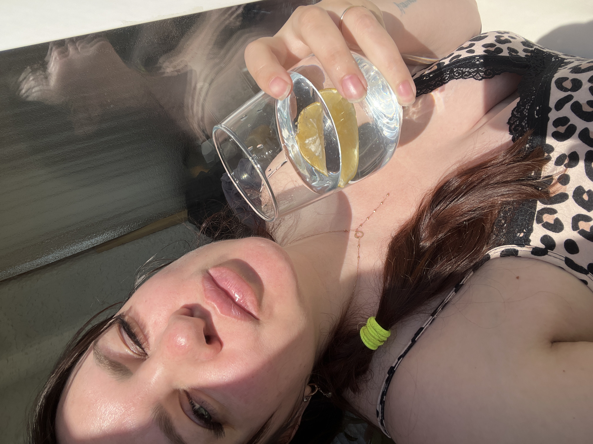A woman is lying down holding a glass of water with a lemon slice in it. She is wearing a leopard print top with black lace trim. She has brown hair pulled back with a neon green hair tie. She is wearing a ring on her right hand and a delicate necklace. The background shows a reflection in what appears to be a magazine cover. The location is unknown, and the city of Noviy, Russia is not discernible.