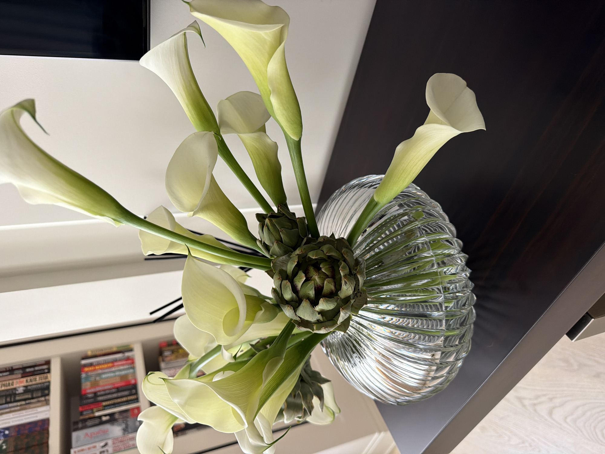 A close-up, eye-level shot captures a bouquet of white calla lilies and artichokes arranged in a fluted glass vase. The vase sits on a dark, polished wooden surface, which appears to be the top of a shelf or table. In the background, a white wall is visible, with a portion of a dark rectangular object, possibly a television, on the right. To the left of the wall, a bookshelf filled with books is partially visible. The lighting is bright, highlighting the delicate petals of the lilies and the textured surface of the vase and artichokes. The overall scene suggests a home or office environment, with a focus on the floral arrangement.