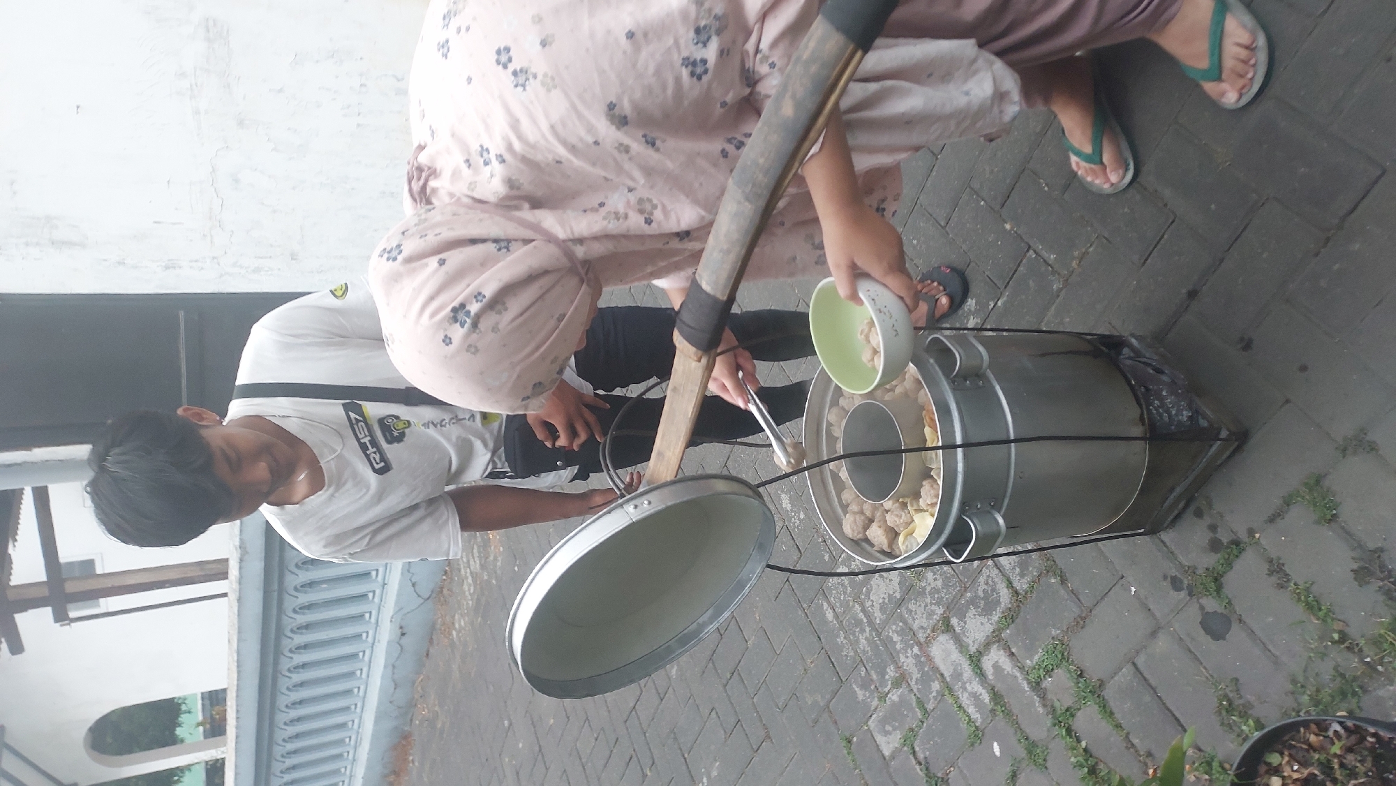 In Malang, Indonesia, two individuals are engaged in preparing food outdoors. A young man wearing a white t-shirt stands behind a large metal pot, which is likely used for cooking. A woman, dressed in a light-colored hijab and patterned clothing, is actively serving food. She is using tongs to place meatballs from the pot into a small green bowl, which she holds in her other hand. The scene unfolds on a paved ground, with some greenery peeking through the gaps between the paving stones. A decorative blue and white fence is visible in the background, suggesting a residential or public area. The overall impression is of a casual outdoor food preparation scene in a community setting.