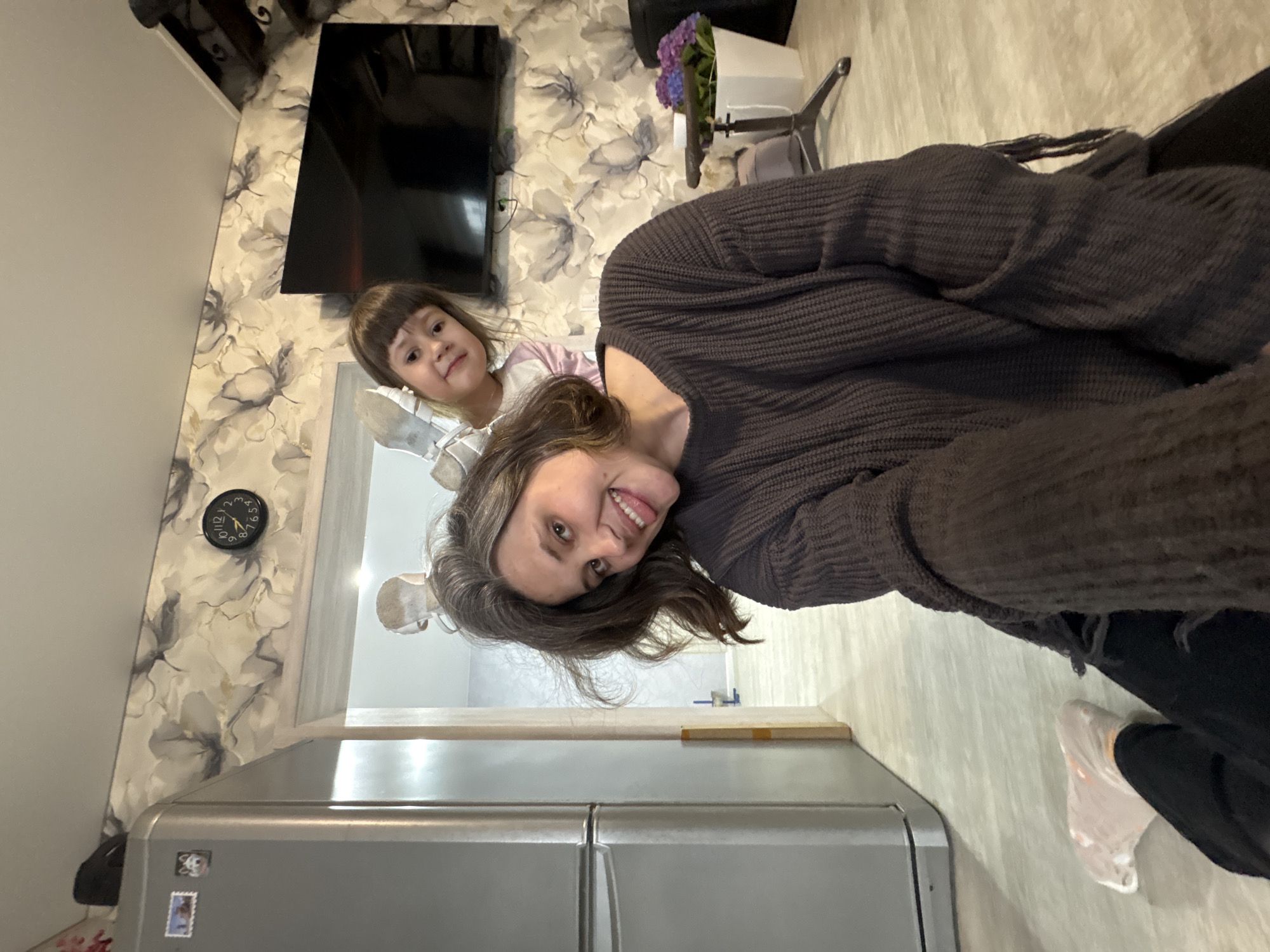 The image shows an indoor scene, likely a residence, in Прикамский, Russia. A woman and a young girl are posing for the camera. The woman is wearing a brown sweater and dark pants. She is smiling with her tongue sticking out. The girl is wearing a light-colored shirt.
Behind them is a wall with floral wallpaper. A flat-screen TV is mounted on the wall. A clock is visible, showing the time as approximately 11:15. A silver refrigerator is in the foreground. A doorway leads to another room.