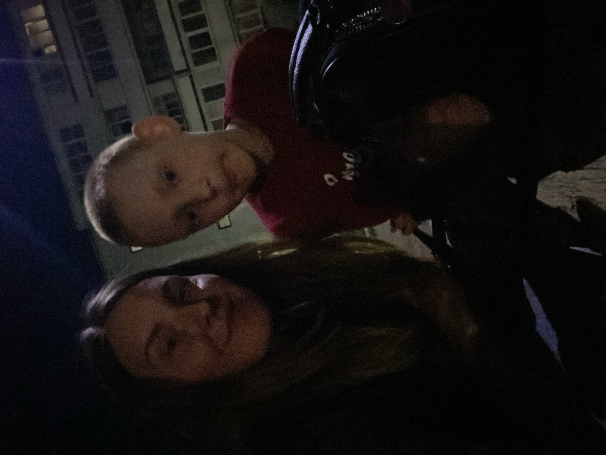 The image, taken at night, shows two people, a woman and a young boy, posing for a selfie. The woman is on the left, with long, dark hair and a light complexion. The boy, on the right, has short, light-colored hair and a fair complexion. He is wearing a red t-shirt. In the background, a multi-story building with illuminated windows is visible, suggesting an urban environment. The overall lighting suggests the photo was taken outdoors in a city location, possibly Noviy, Russia, given the context. The image quality is grainy, likely due to low light conditions.
