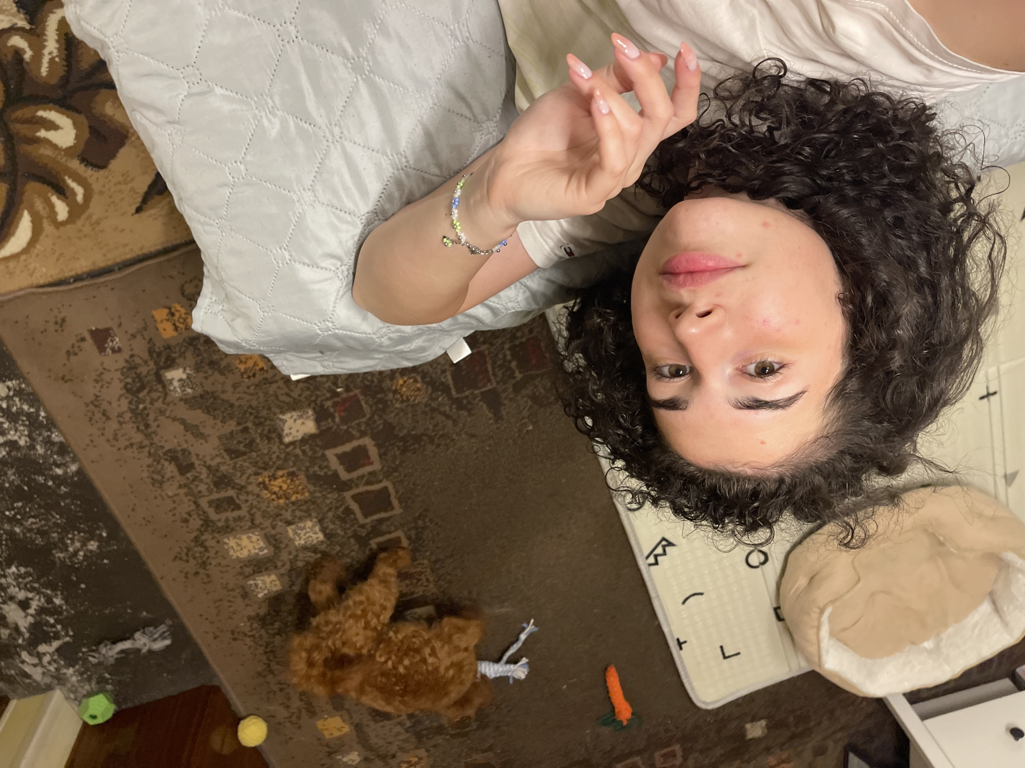 The image captures a high-angle, indoor scene likely within a residential setting. A person with dark, curly hair and light skin is lying down, their face partially visible in the foreground. They are wearing a light-colored t-shirt. A light blue, quilted blanket is positioned beside them. In the background, a small, reddish-brown dog is lying on a patterned rug. Various toys are scattered on the rug, including a green ball, a rope toy, and a toy carrot. The overall impression is one of a casual, relaxed domestic environment. The location in the city of Sofia, Bulgaria is not discernible from the image itself.