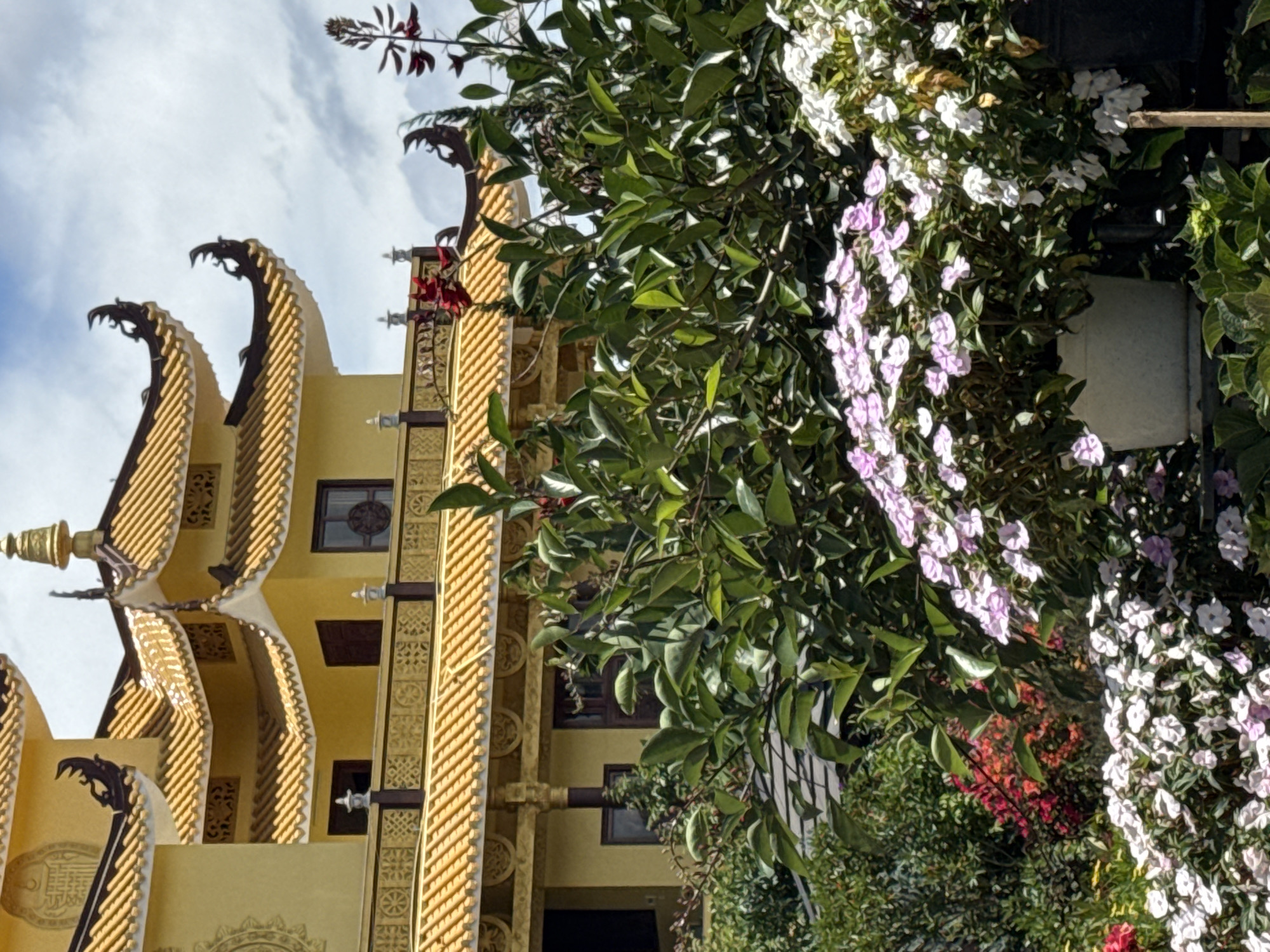 The image depicts a portion of a yellow building with multiple tiered, orange-tiled roofs, characteristic of Buddhist temple architecture. The roofs have decorative upturned eaves adorned with dark, carved figures. Ornamental railings with intricate patterns run along balconies. In the foreground, a lush garden overflows with greenery and a variety of flowering plants, including clusters of small pink and white flowers in a white pot, and scattered red and dark red blossoms. The backdrop consists of a cloudy sky. This architectural style and lush garden setting are consistent with the Truc Lam Zen Monastery, a prominent Buddhist monastery located on a hill overlooking Xuan Huong Lake in Da Lat, Vietnam.