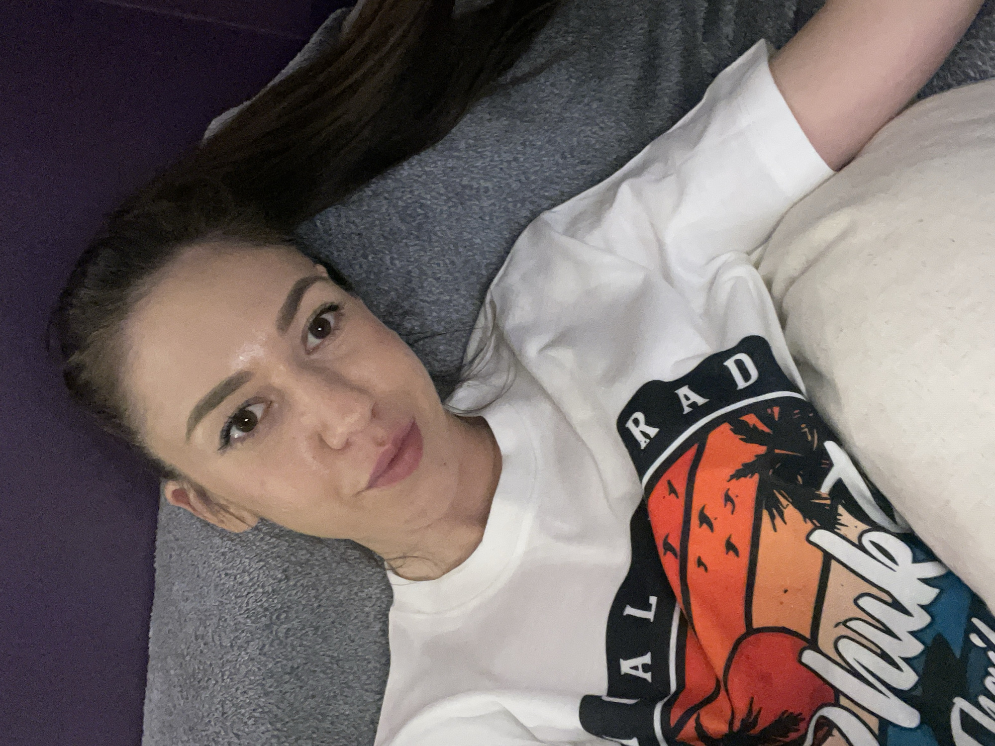 A woman lies on a grey surface, possibly a couch or bed, with a white pillow to her right. She is wearing a white long-sleeved shirt with a colorful graphic design that includes palm trees and the word "RAD" in black letters. Her dark hair is pulled back, and she is looking towards the camera. The background is a deep purple color, possibly a wall or another piece of furniture. There is no discernible information to identify the location as Sofia, Bulgaria.
