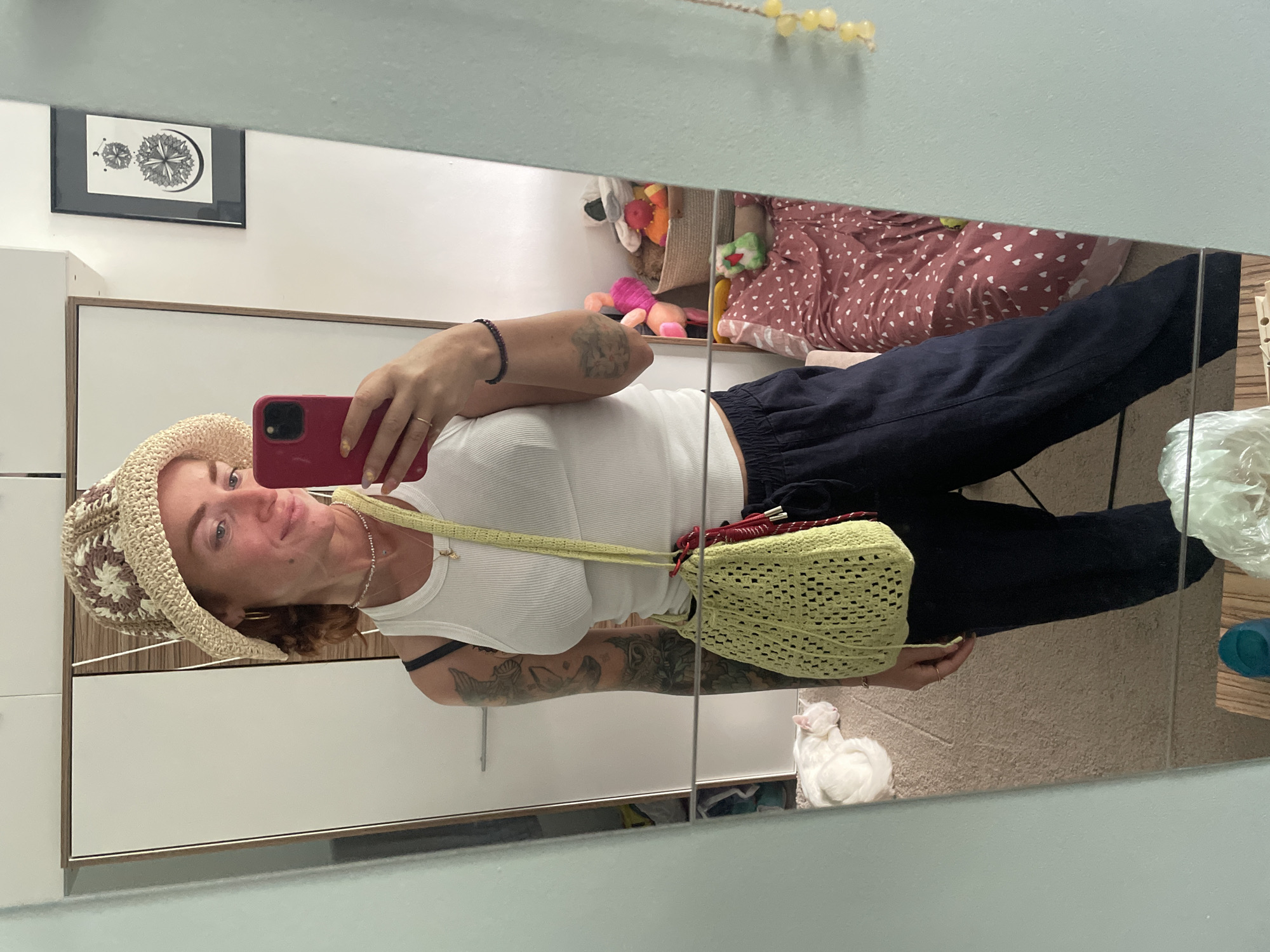 A woman stands in front of a full-length mirror, taking a selfie with her phone. She is wearing a white ribbed tank top, dark blue drawstring pants, and a crocheted bucket hat with a floral pattern. A light green crocheted bag hangs from her shoulder, with a red drawstring. She has a delicate necklace and a beaded bracelet on her wrist. Tattoos are visible on her arms. In the reflection of the mirror, a white cat is sleeping on a beige carpet. Behind the cat, a basket filled with stuffed animals is visible, along with a pink and white patterned blanket. On the wall behind the cat, a framed black and white drawing is hanging. The room appears to be a bedroom. The lighting suggests it is daytime.