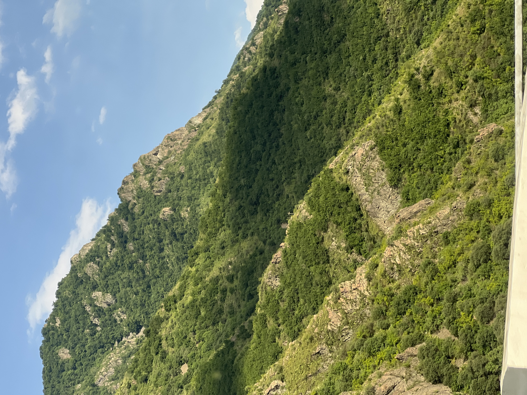 The image shows a steep, verdant mountainside in the vicinity of Alaverdi, Armenia. The mountain is covered with dense green foliage, with rocky outcrops breaking through the vegetation. A large shadow falls across the middle section of the mountain, indicating the sun is positioned to the left. Two small, white crosses are visible on the slope, suggesting a religious or memorial site. The sky above is a clear blue with a few scattered white clouds. In the foreground, a portion of a white concrete barrier is visible, implying the photo was taken from a road or elevated viewpoint. The overall scene is one of natural beauty and rugged terrain.