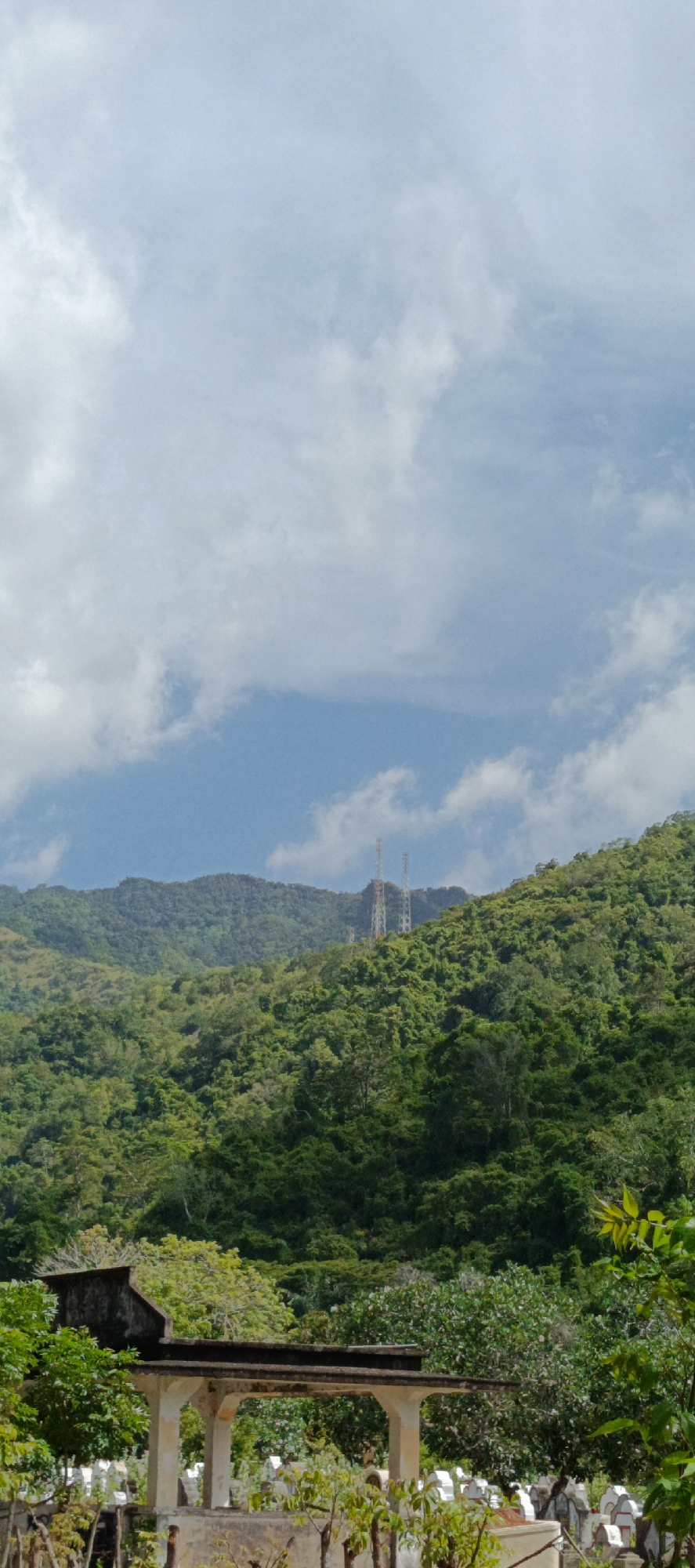 A vertical, medium shot captures a lush, green mountainside in Geundring, Indonesia, under a partially cloudy sky. In the foreground, a weathered, open-air structure with pillars and a flat roof sits in front of a cemetery with numerous white tombstones. The middle ground is dominated by dense, vibrant green foliage covering the sloping terrain. Two tall, lattice towers, possibly for communication or broadcasting, are visible in the distance, perched on the crest of the mountain range. The sky above is a pale blue, with soft, white clouds scattered across it. The image appears to be taken during daylight hours.