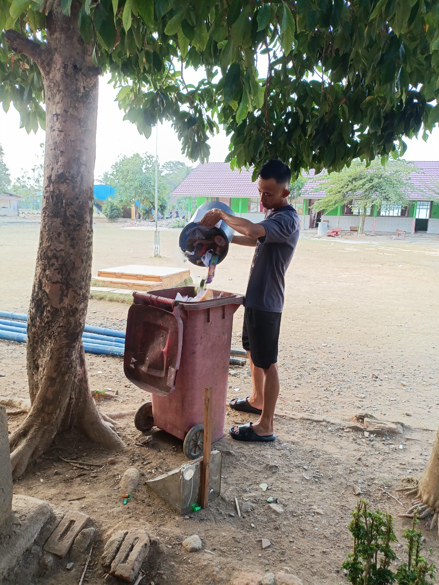 A man is shown in the city of Sumberejo, Indonesia, standing next to a large, weathered maroon trash bin. He is wearing a dark gray t-shirt and black shorts, with black sandals on his feet. The man is in the process of emptying a smaller dark blue bucket into the larger trash bin. The bucket appears to contain a variety of discarded items, including what look like food wrappers and plastic. The scene is outdoors, with a large tree providing shade over the man and the trash bin. In the background, there are buildings with red roofs, suggesting a school or community area. The ground is covered with dirt and small stones. There are also some blue pipes lying on the ground to the left of the trash bin.