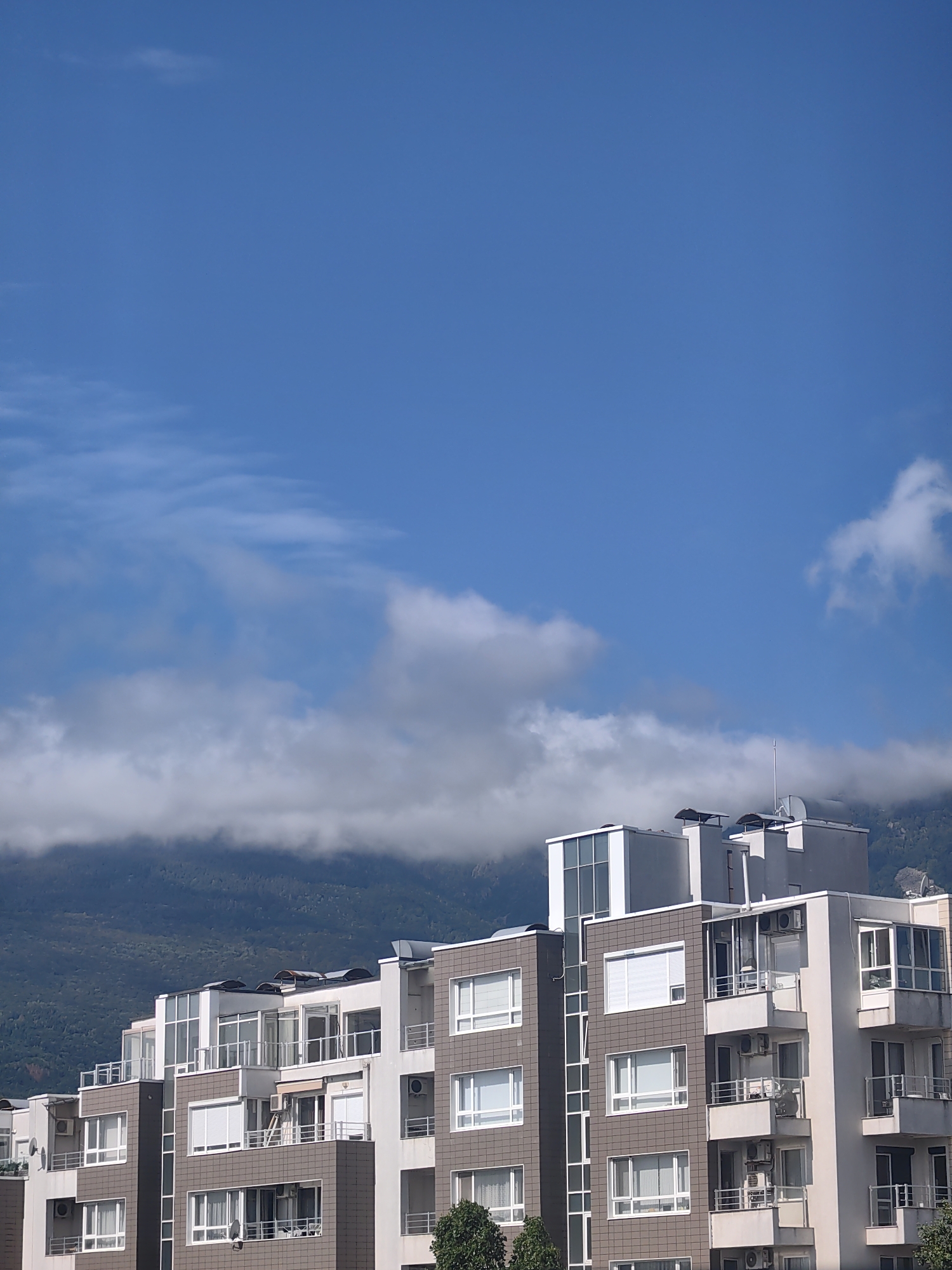 The image shows a modern apartment building with multiple floors, balconies, and windows against a blue sky with scattered clouds. The building has a contemporary design with sections of brown tiled facade and white walls. The top of the building features several air conditioning units, satellite dishes, and ventilation systems. In the background, a forested mountain range is visible, partially obscured by clouds. The scene appears to be in a city setting, likely Sofia, Bulgaria, given the architecture and mountainous backdrop often seen in the city's outskirts. There are no people visible in the image. The overall impression is one of urban residential development situated near a natural landscape.