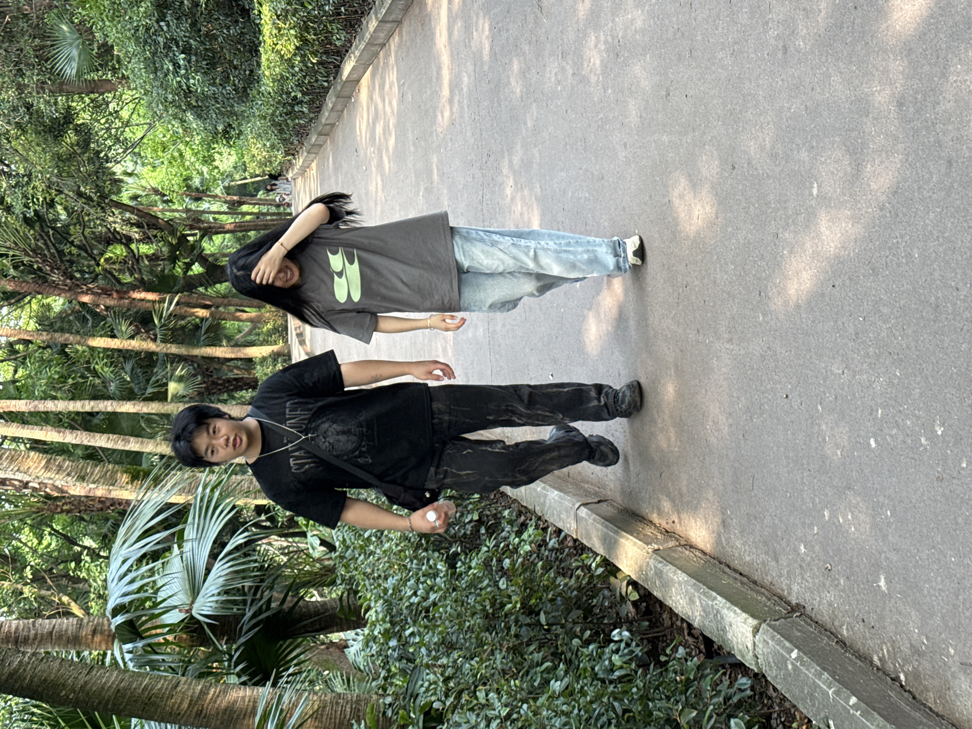 Two individuals are positioned on a paved pathway within a park setting. The male subject, attired in a black shirt and dark pants, is facing the camera. The female subject, dressed in a gray shirt and blue jeans, partially obscures her face with her hand. The surroundings are characterized by dense foliage, including trees and shrubs. The image was taken in the Jiulongpo District of China.