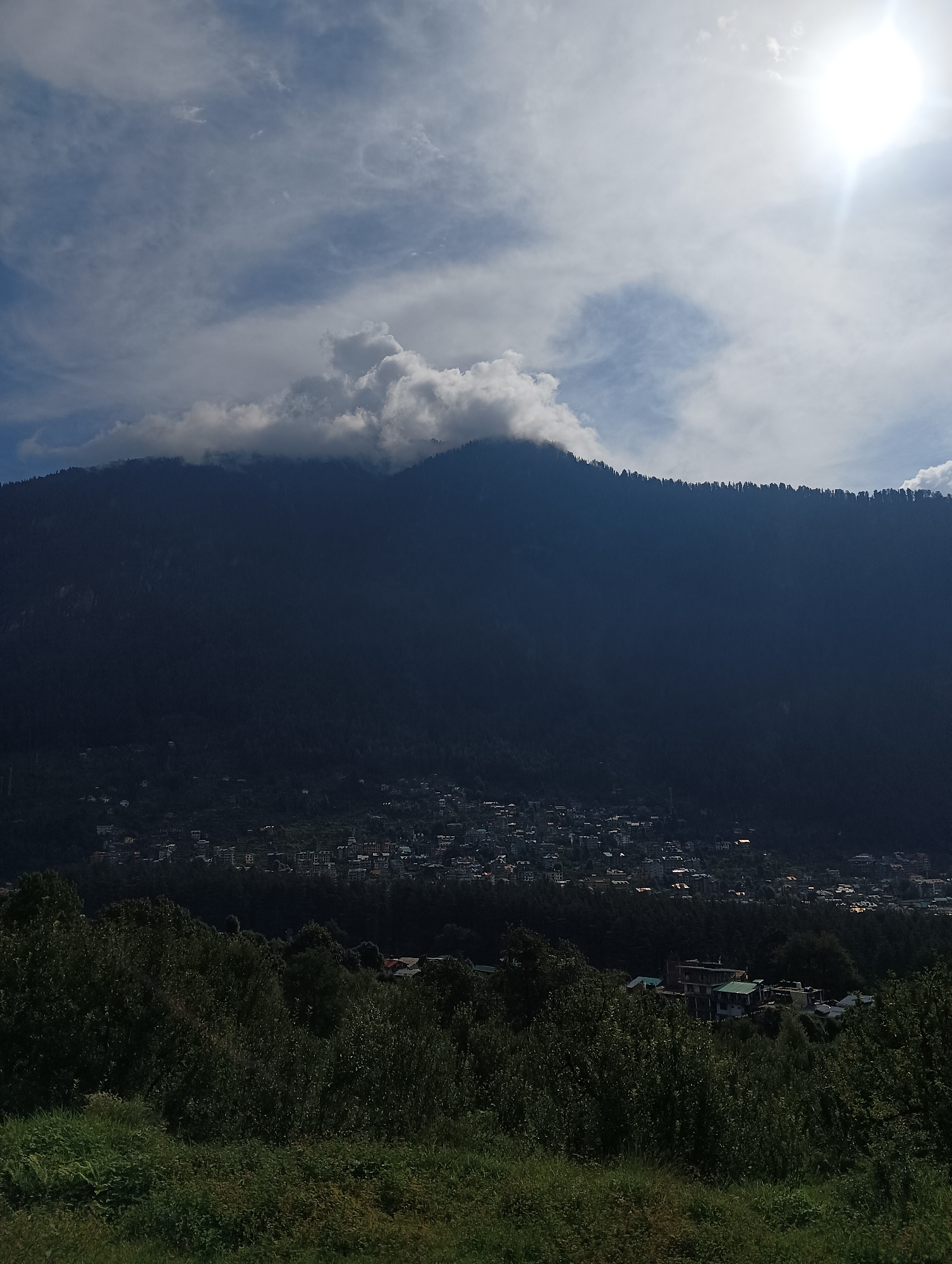 The image captures a panoramic view of Manali, India, from an elevated vantage point. In the foreground, lush green vegetation blankets the lower portion of the frame. A dense forest, comprising numerous trees and shrubs, extends upwards, partially obscuring a view of the town nestled at the base of a mountain.
The town of Manali itself is visible as a cluster of buildings, with houses and other structures spread across the hillside. The architecture appears to be a mix of traditional and modern styles. Some buildings have red or tiled roofs, while others are painted in various colors.
Dominating the midground and background is a colossal, dark mountain, its slopes covered in a dense canopy of trees that form a sharp, dark line against the lighter sky. A band of fluffy white clouds clings to the upper portion of the mountain, creating a dramatic contrast with the dark massif.
The sky above is a brilliant blue, partially obscured by wispy white clouds. A bright sun shines from the upper right corner of the image, casting a strong glare and illuminating the clouds. The overall lighting suggests it is daytime, likely late morning or early afternoon, with the sun at a relatively high angle. The scene evokes a sense of natural grandeur and the peaceful coexistence of human settlement with a majestic natural landscape.