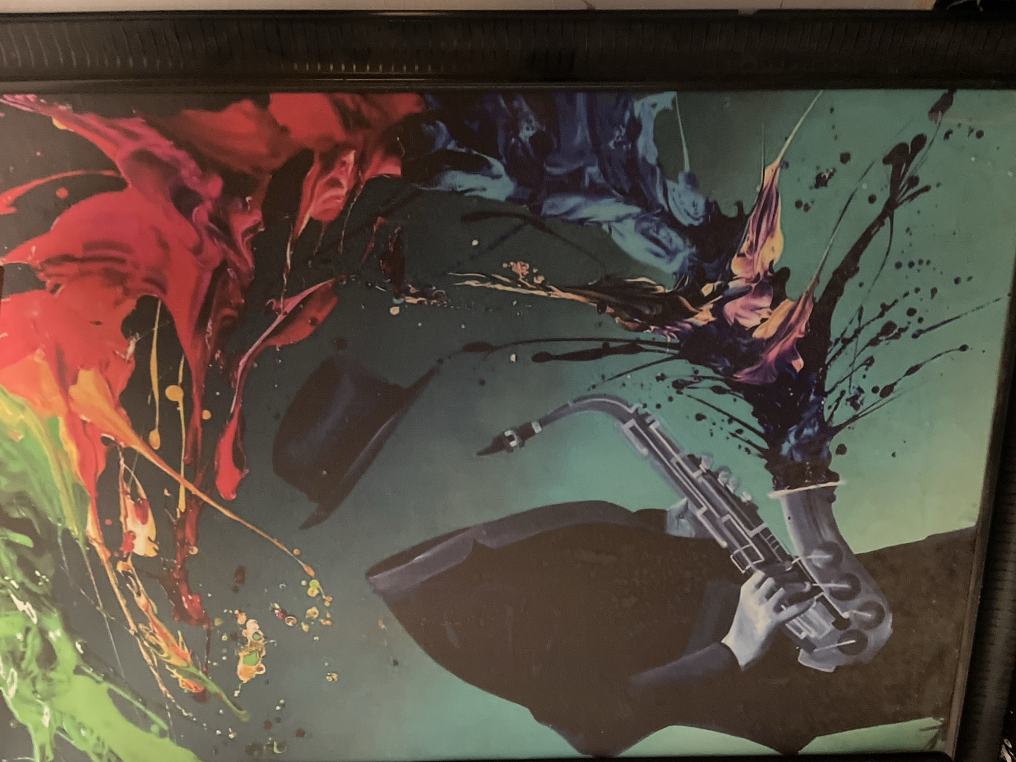 This image is a dynamic and colorful painting that combines figurative and abstract elements. The central subject is a stylized figure of a musician, depicted in a dark silhouette with subtle highlights, playing a saxophone. The musician's hands are visible holding the instrument, which also has some visible details like keys and reflective surfaces. A dark fedora hat floats seemingly above the musician to the left.
The background is a rich, dark teal or deep green color. Surrounding the musician and extending across much of the canvas are vibrant, abstract splatters and drips of paint. These bursts of color include vivid reds, bright greens, deep blues, and hints of yellow and purple, creating a sense of explosive energy and movement. The paint appears to emanate from different points, suggesting the dynamism and passion of a musical performance.
The overall scene conveys a powerful artistic expression, likely representing the lively essence of jazz or music itself. The blend of fluid abstract forms with the distinct musical subjects evokes a strong sense of rhythm and emotion. There is no visible text or specific location cues within the artwork itself. The image does not provide any specific time of day or weather details, focusing instead on the artistic concept.