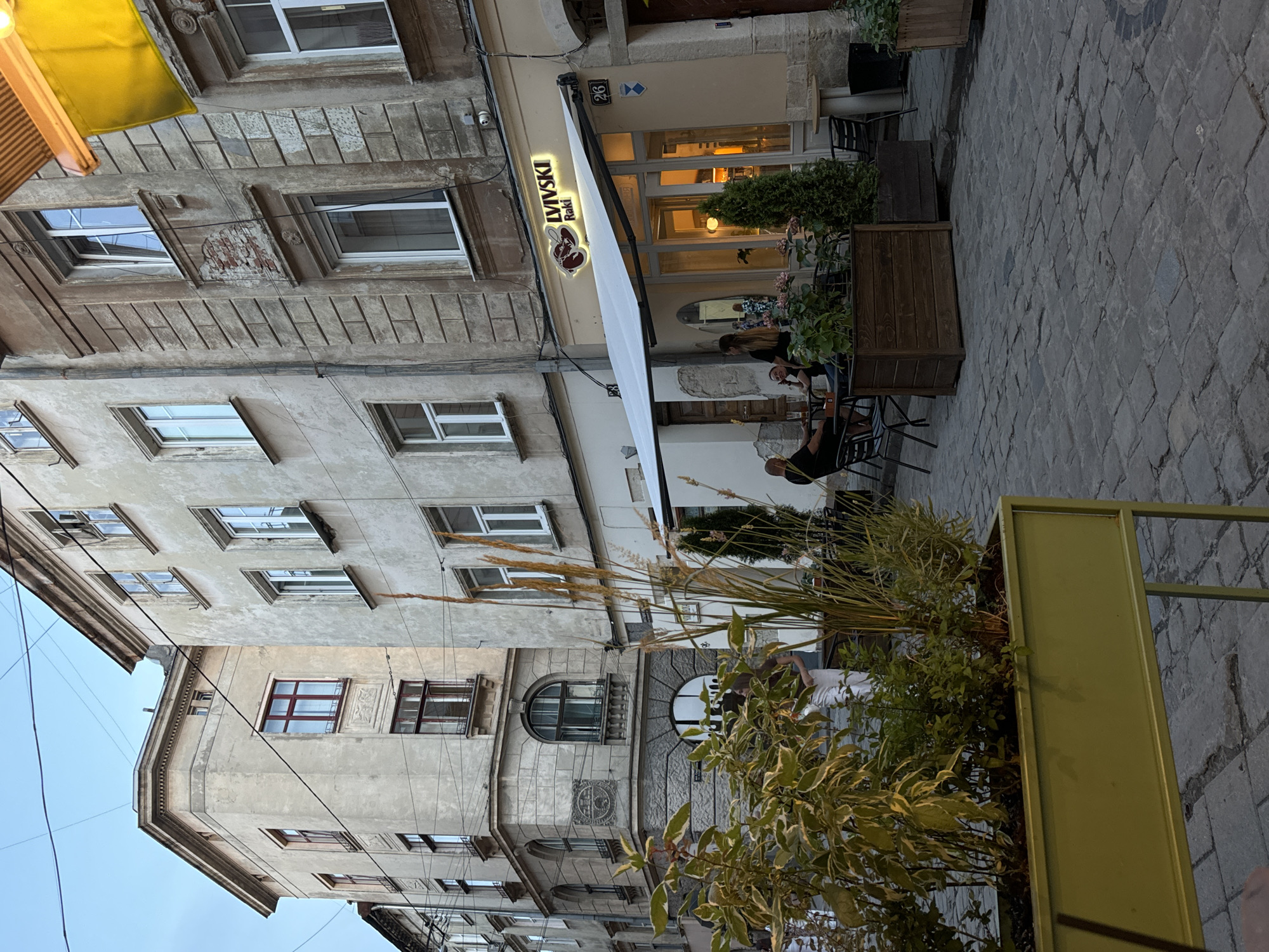 This media file captures an outdoor seating area of a cafe or restaurant on a cobblestone street. The scene is viewed from a low angle, looking up towards the facades of several old buildings. The architecture suggests a European city, with intricate stonework and weathered plaster walls. A bright yellow awning is visible on the left, and a white retractable awning extends over the outdoor seating area of the establishment.
The cafe's signage, illuminated with a neon-like glow, reads "LVIVSKI Raki". It features a stylized logo with a red shape. Several people are seated at small tables, some engaged in conversation, others looking at their phones. The atmosphere appears relaxed and casual. Two large wooden planters filled with greenery and tall ornamental grasses add a natural element to the urban setting. A green metal planter box is prominently featured in the foreground.
The time of day appears to be late afternoon or early evening, as the light is soft and shadows are beginning to lengthen. There are no strong indications of specific weather conditions, but the sky appears overcast or the lighting is subdued. The overall impression is one of a charming, perhaps historic, urban environment where people are enjoying an evening out. The visible text "LVIVSKI Raki" strongly suggests a location in Lviv, Ukraine, known for its Raki (a type of brandy).