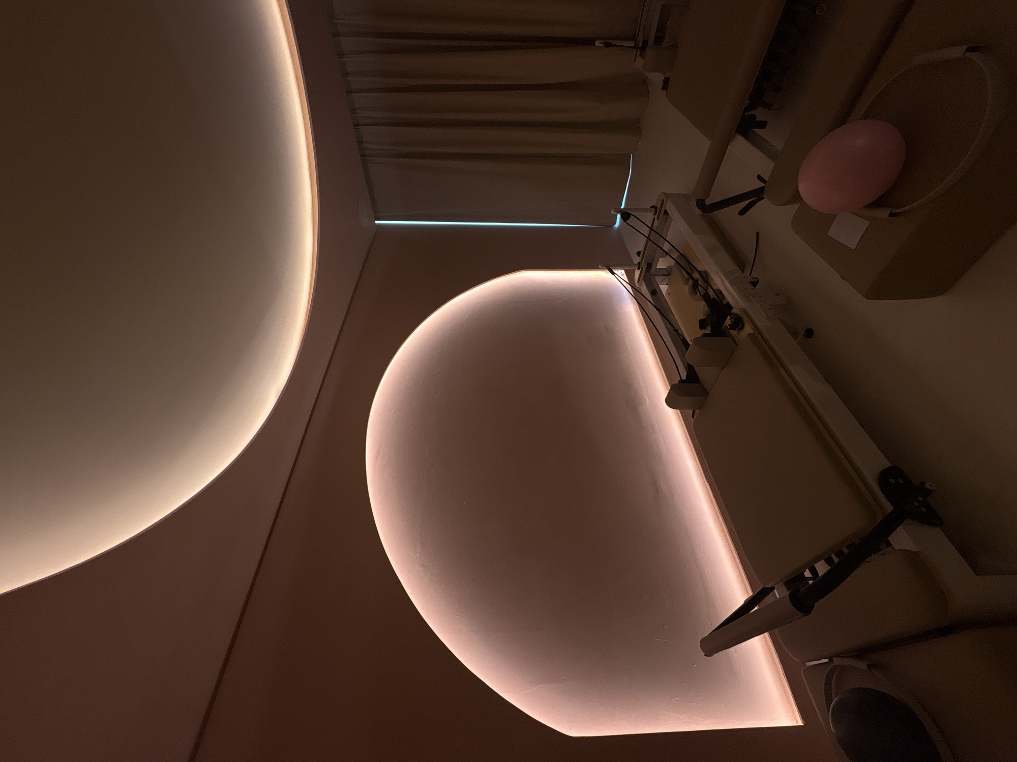The image depicts an interior space, likely a medical or therapeutic setting, possibly in Mexico City, Mexico, given the request to provide the location. Dominating the scene are large, curved, illuminated architectural features, possibly wall or ceiling designs with embedded lighting. A medical examination table with various adjustable components is visible, with a pink, possibly latex, balloon or cushion placed on the table. Curtains are visible in the background. The lighting in the room is dim, creating a warm, somewhat clinical ambiance. No people are visible.