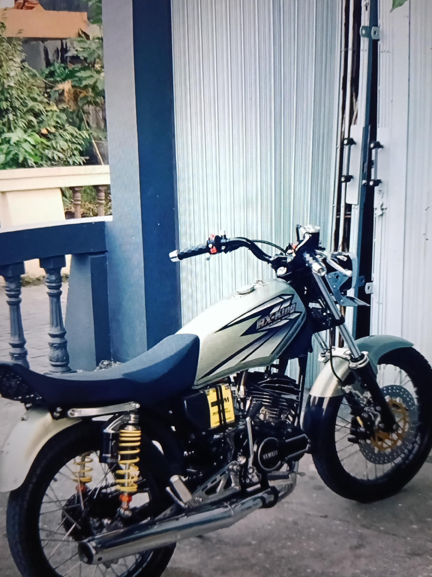 The image shows a light-colored Yamaha RX-King motorcycle parked on a concrete surface in front of a grey corrugated metal gate. The motorcycle is positioned diagonally, with its front wheel closer to the viewer. It features a dark blue seat, yellow rear shock absorbers with red accents, and a chrome exhaust pipe. The motorcycle's engine is visible, and the "YAMAHA" logo is present on the engine casing.
In the background, to the left of the motorcycle, is a dark blue pillar and a railing with decorative posts. Beyond that, there are green trees and a building with a beige facade. The overall setting appears to be an outdoor area, possibly a driveway or a yard, in a residential or commercial area within the city of Raha, Indonesia. The lighting suggests it might be daytime, with diffused light. No people are visible in the image.