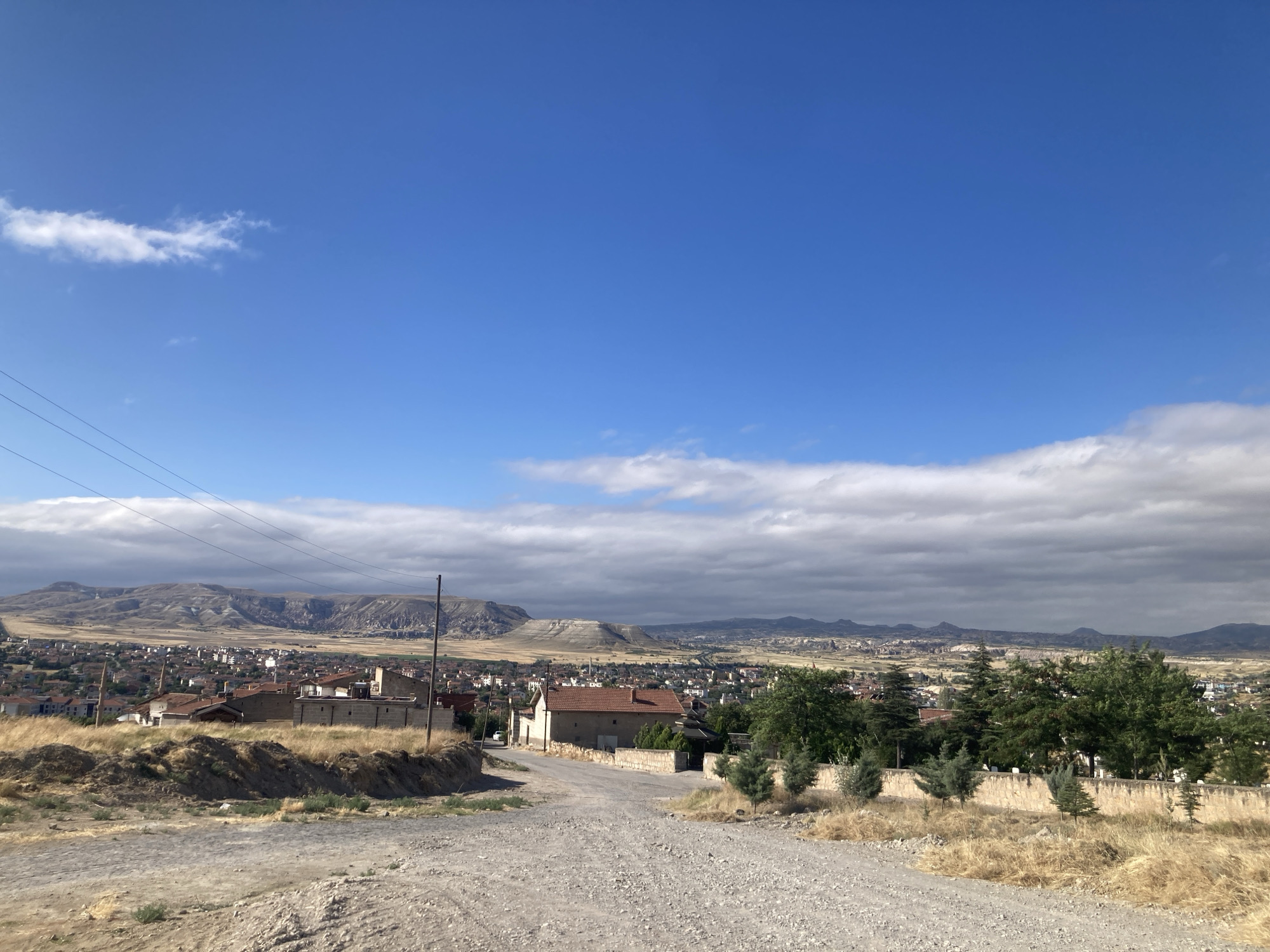 This is a wide-angle, eye-level outdoor shot of a rural landscape on a bright day with a clear blue sky and scattered white clouds. In the foreground, a dirt road curves uphill, flanked by dry, yellow grass. A power line runs horizontally across the top left of the image. In the middle ground, a small town with red-tiled roofs and beige buildings is nestled into the terrain. Beyond the town, a range of dry, rocky hills stretches across the horizon. The overall impression is one of a sunny, tranquil, and sparsely populated area.